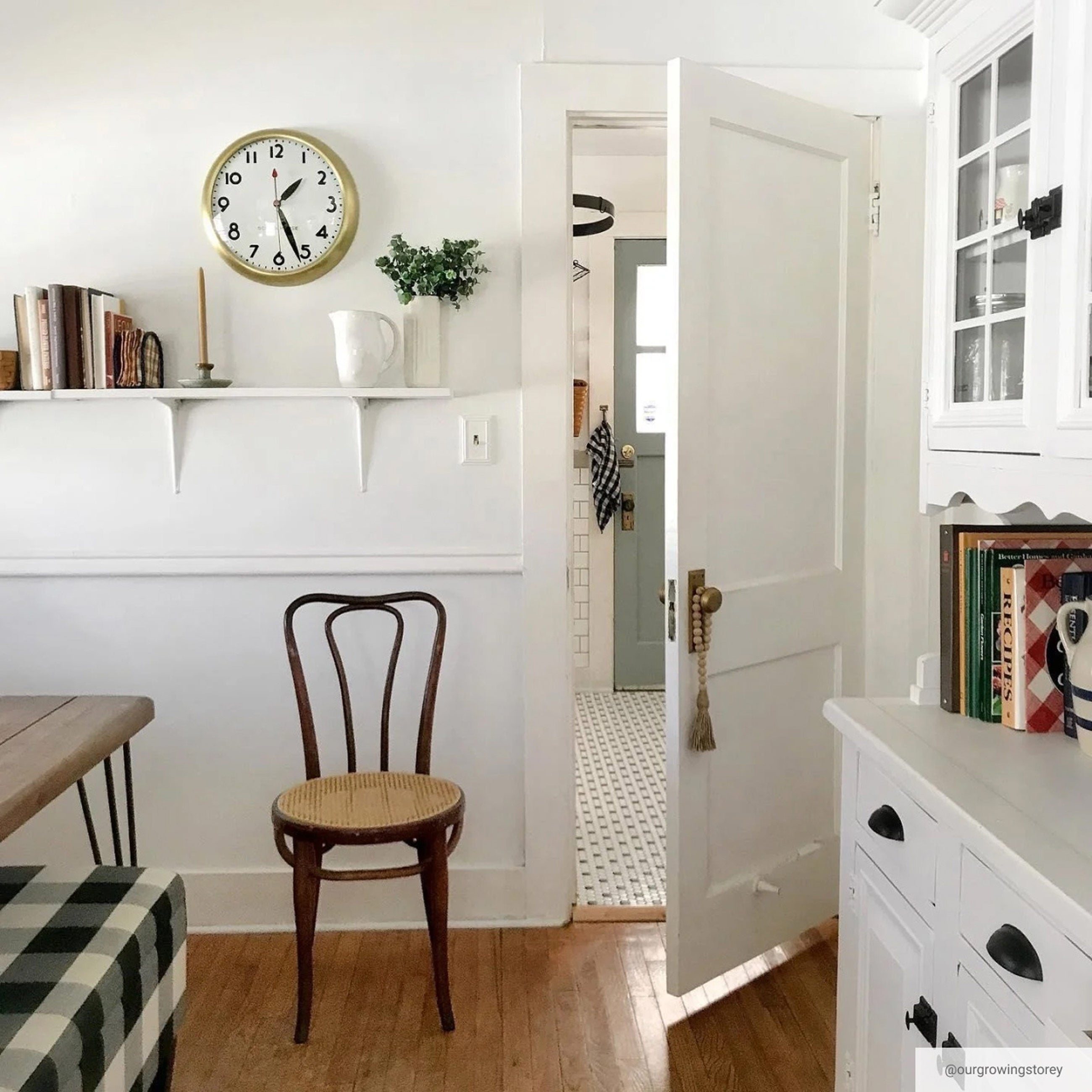 Brass wall clock above white shelf holding books and greenery in bright white kitchen space.