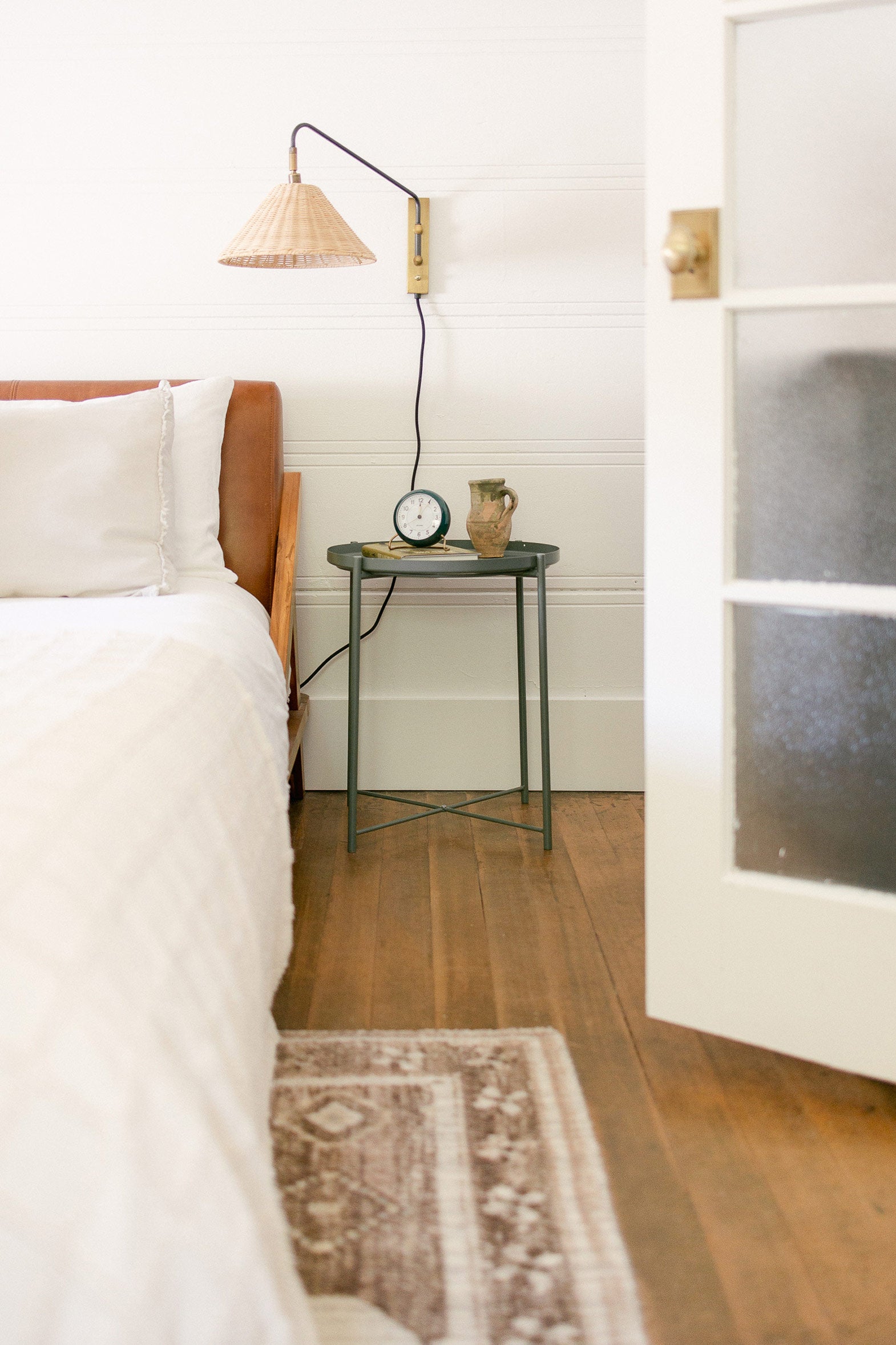 Bright and airy bedroom with small green bedside table holding a tiny clock and vase with a rattan wall sconce above.