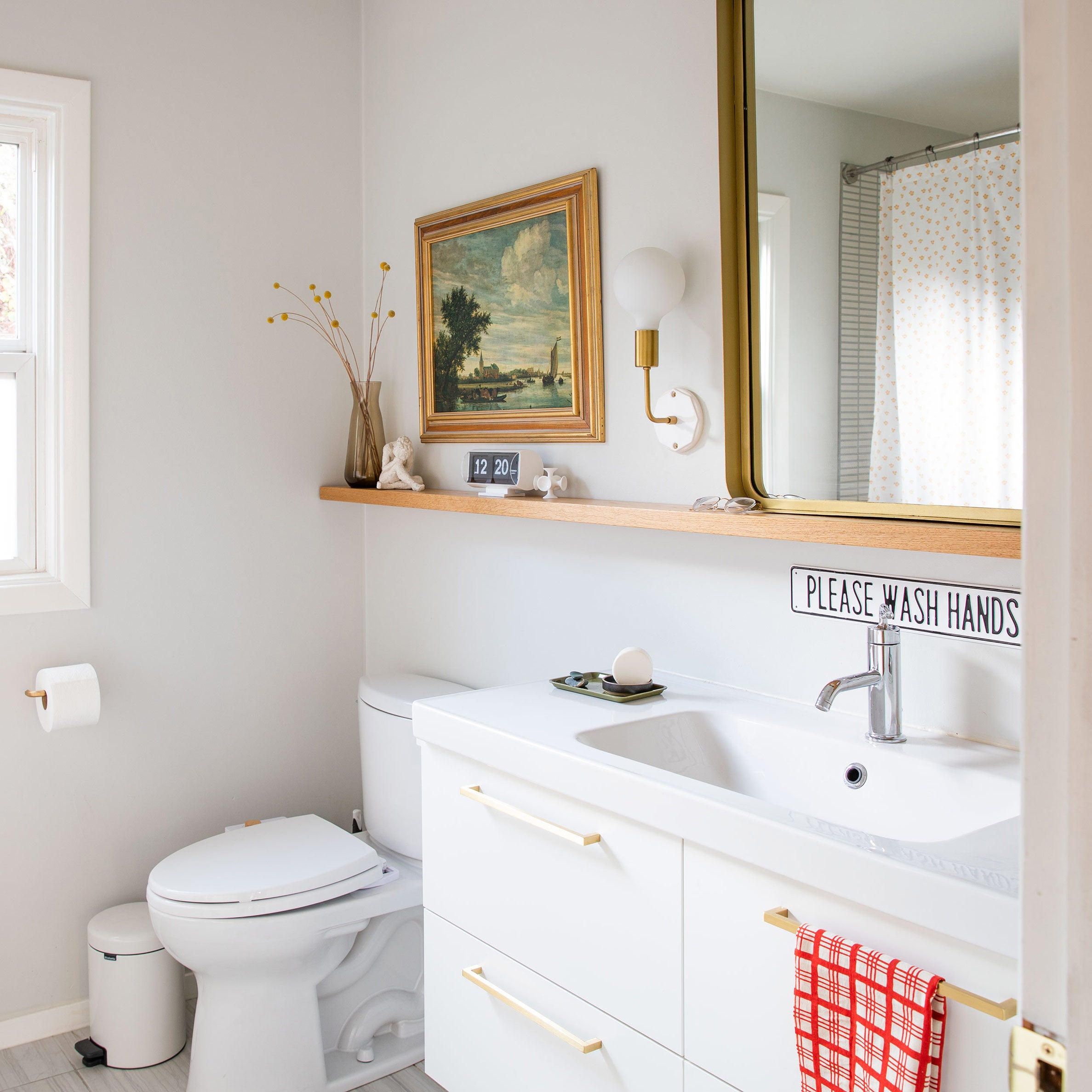 Bright and light bathroom with long shelf above sink and toilet holding clock, flowers in a vase, and a large picture frame.