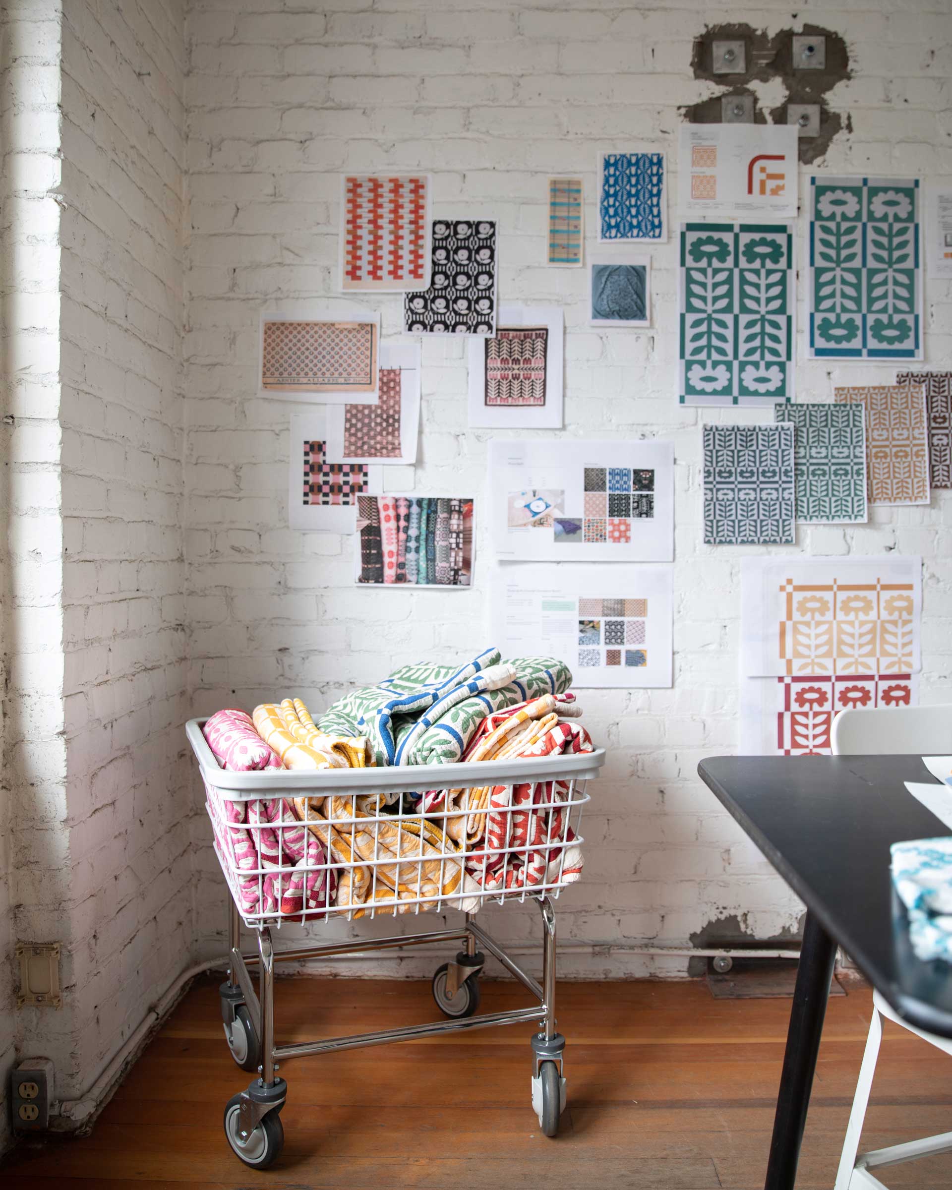 A cart full of Stillwater Floral Quilts in various colors in front of a white brick wall with inspiration images pinned up.