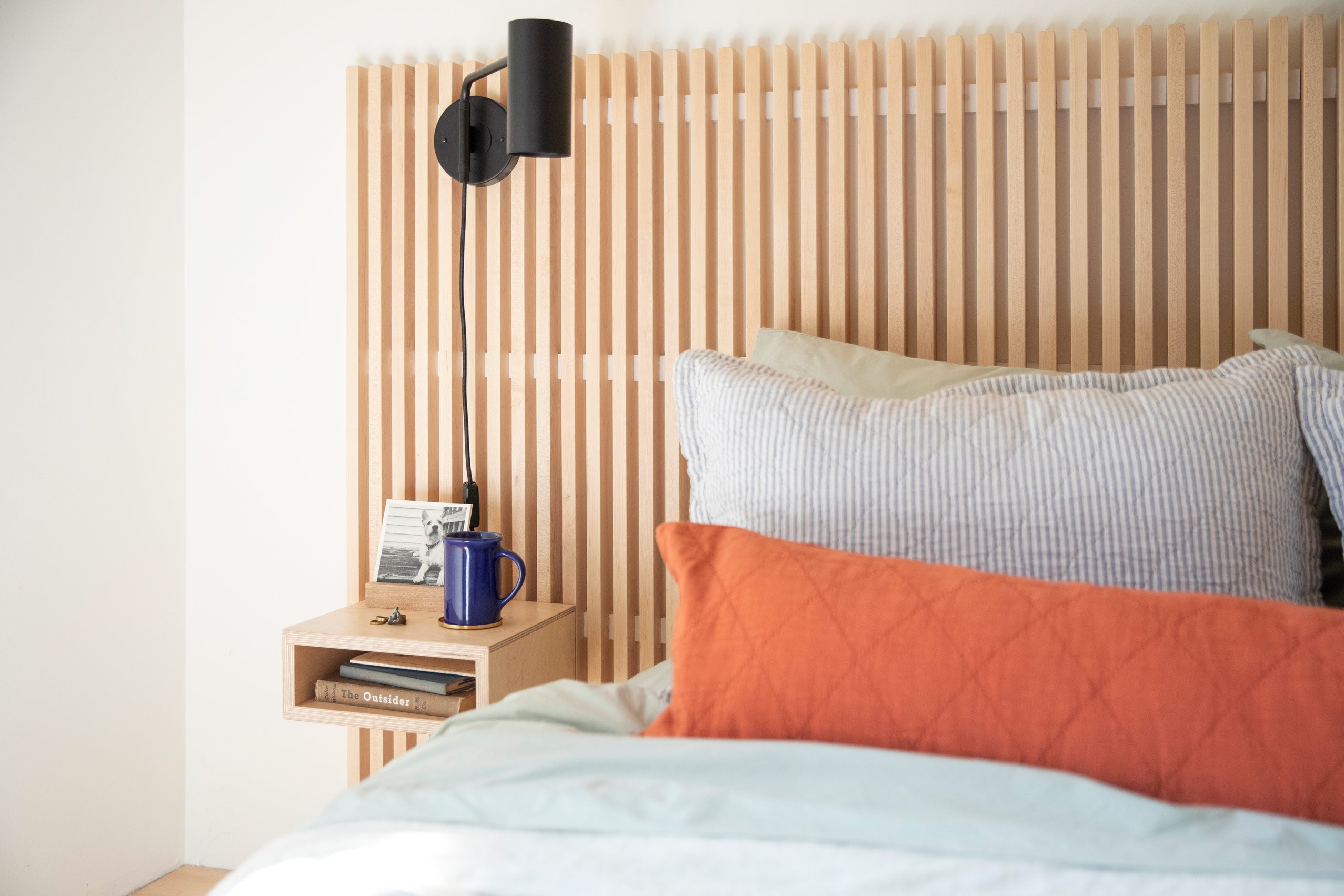 Cozy bed with pinstripe bedding and red bolster pillow and wooden slat wall behind it featuring a black wall sconce and small bedside table with a blue meg and picture frame