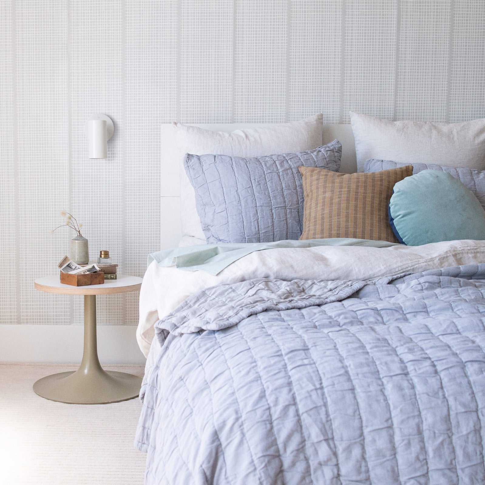 Cozy bed with light blue bedding and bedside table with small bud vase and wooden box containing photos with a white sconce above.