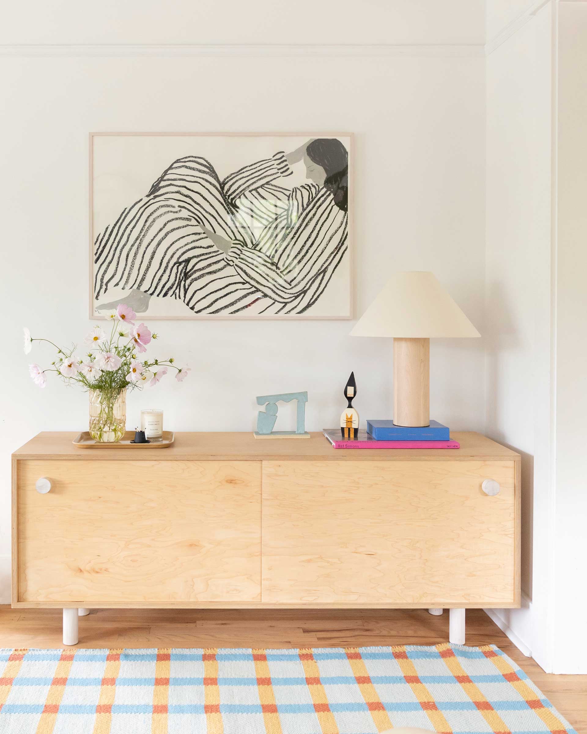 A wood credenza with a wood base table lamp and art piece hanging on the wall.