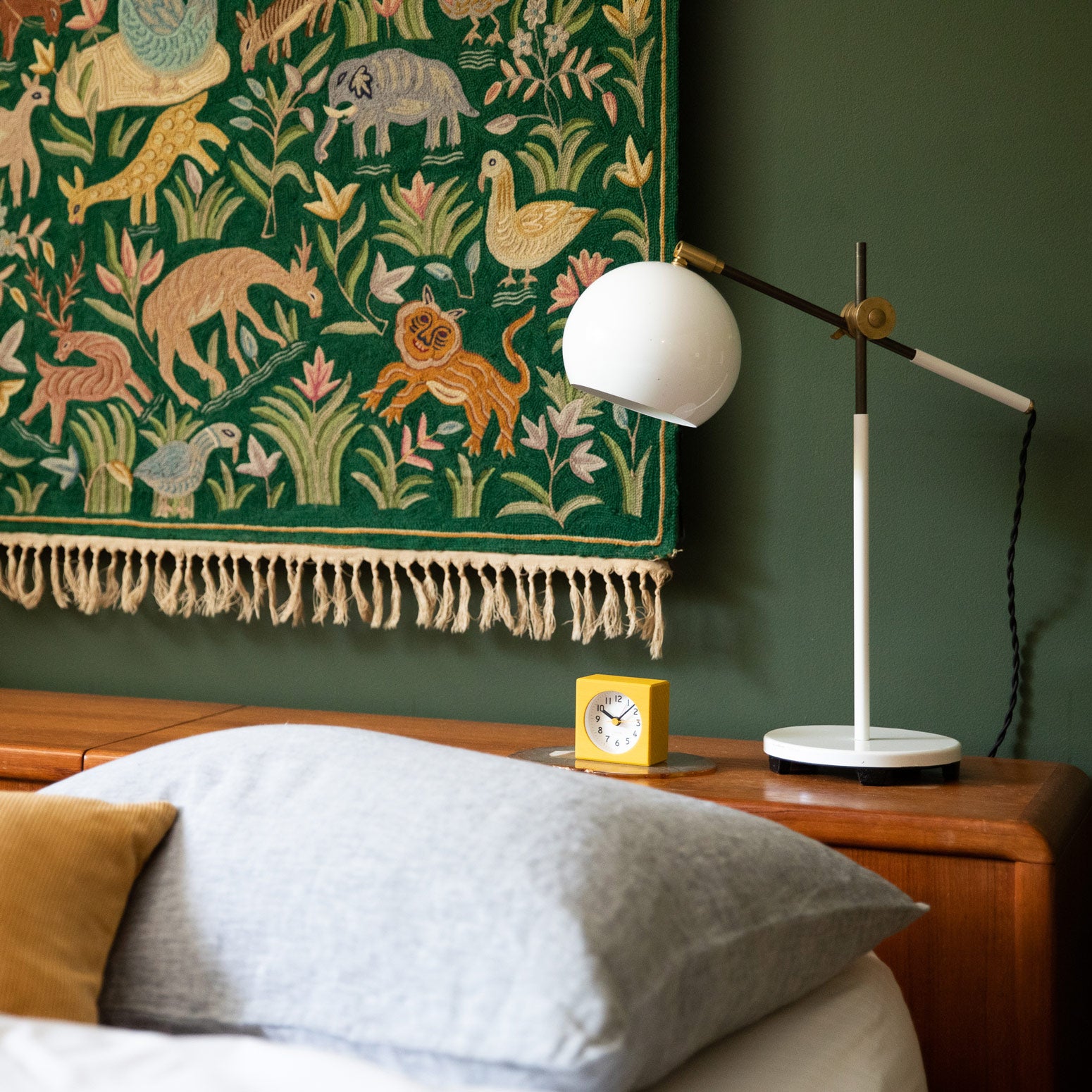 Moody green bedroom with hanging tapestry above the bed and a white table clock and yellow woodblock clock by the bedside.