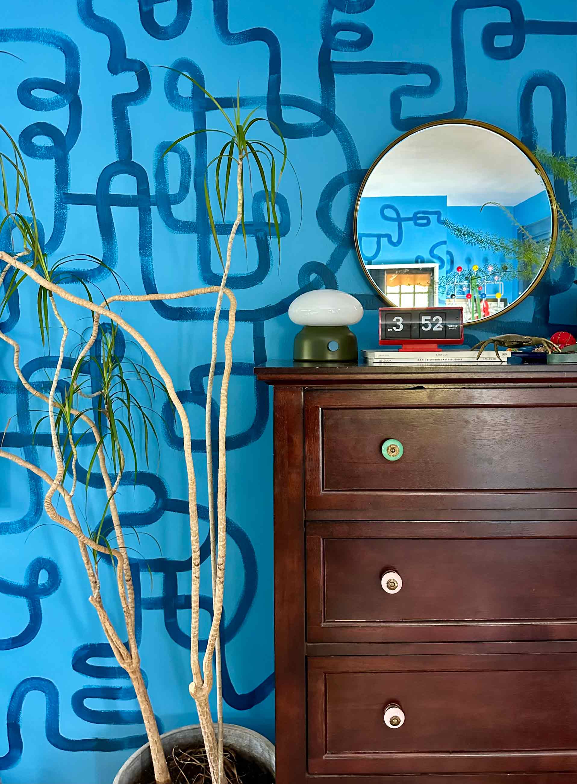 Wooden dresser in bedroom with lamp on top of it and a circle mirror above it.