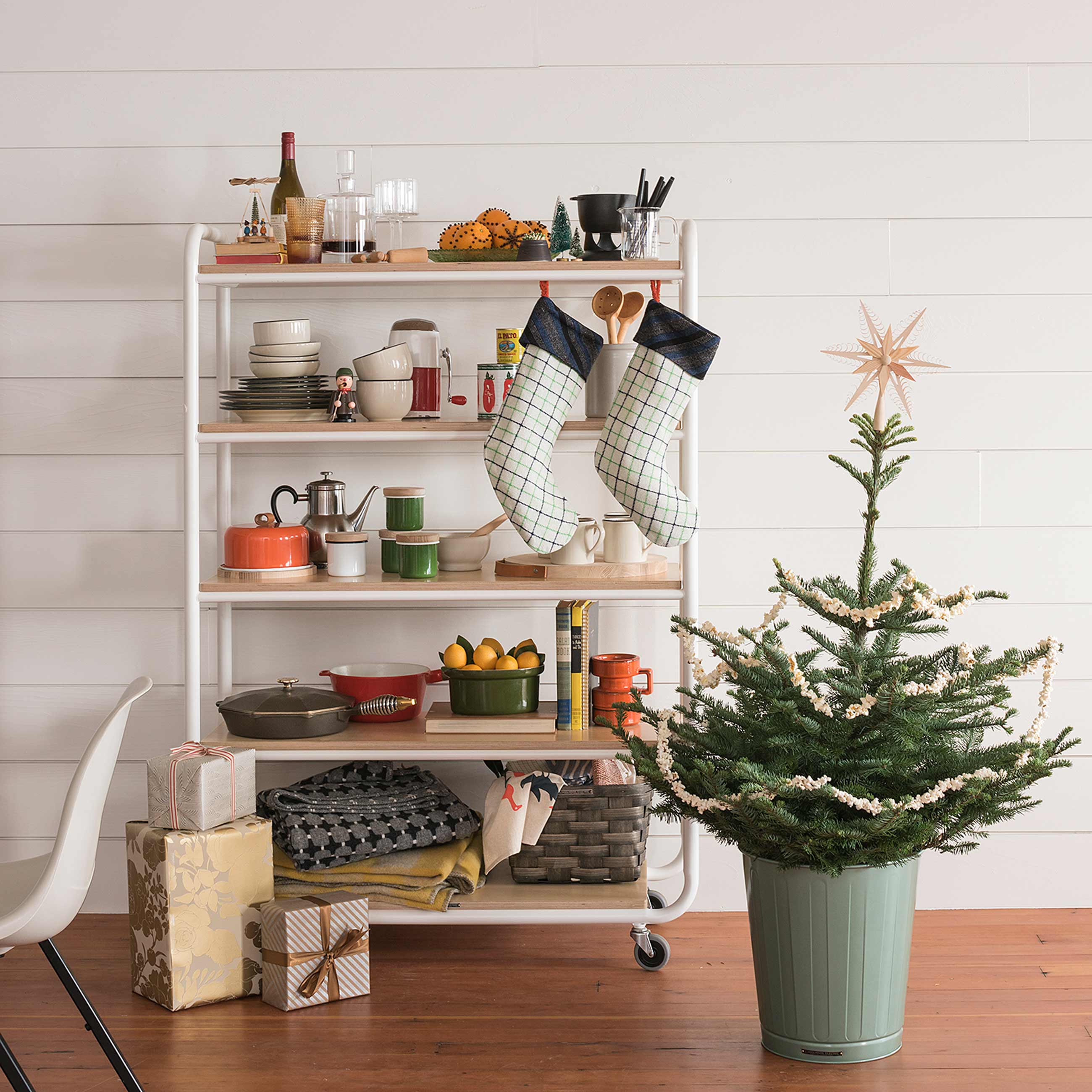 Eclectic kitchen cart with various kitchenwares and two stockings hanging from it next t o small Christmas tree in green pot.