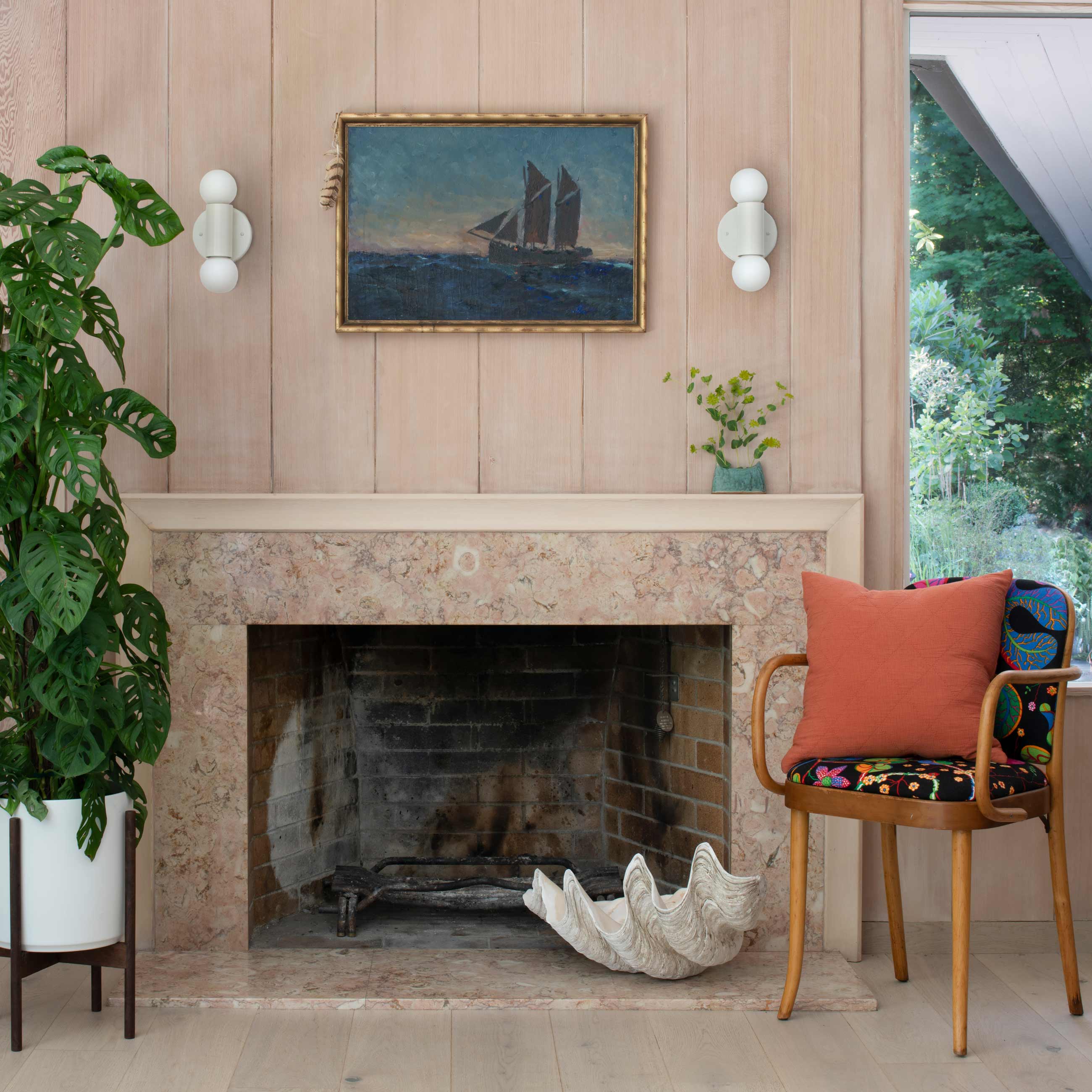 Fireplace with ship painting and two sconces hanging above it as well as a wooden chair and potted plant next to it.