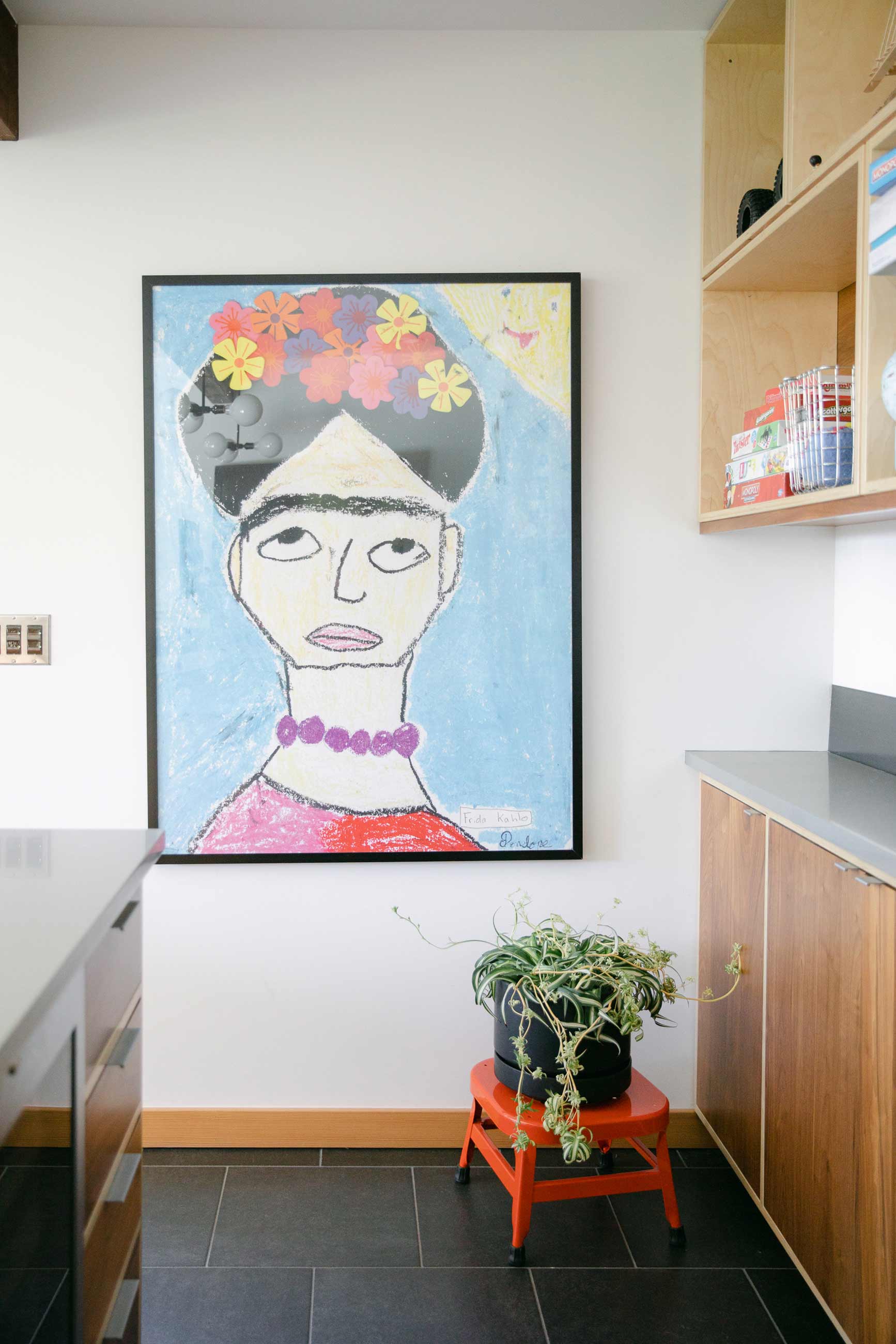 Large scale Frida Kahlo painting in kitchen hanging above small, red utility stool with a thriving potted plant on top of it.