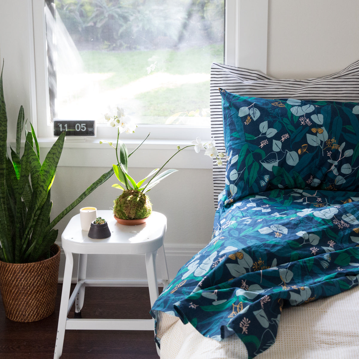 blue and white pillow on a white bed next to a plant on a stool and a window