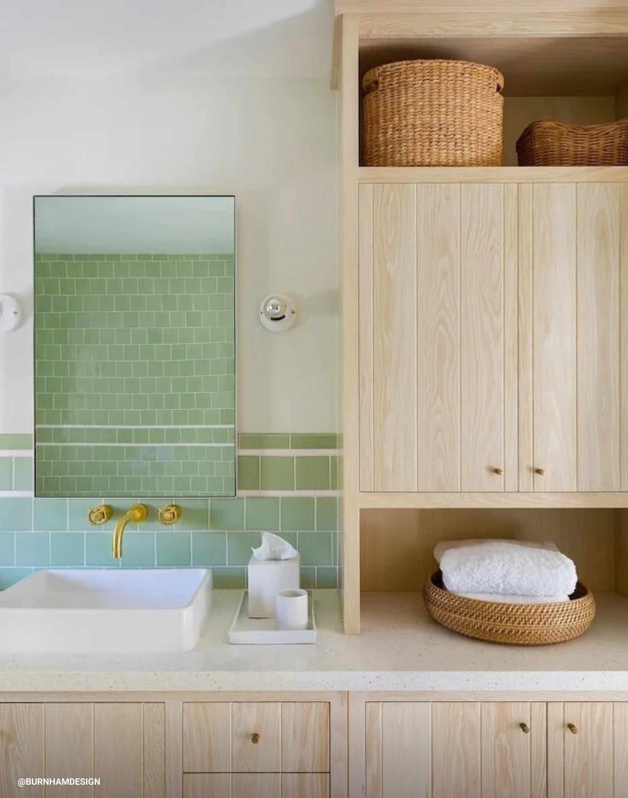 Light wood bathroom cabinets next to white sink with green tile above it. 