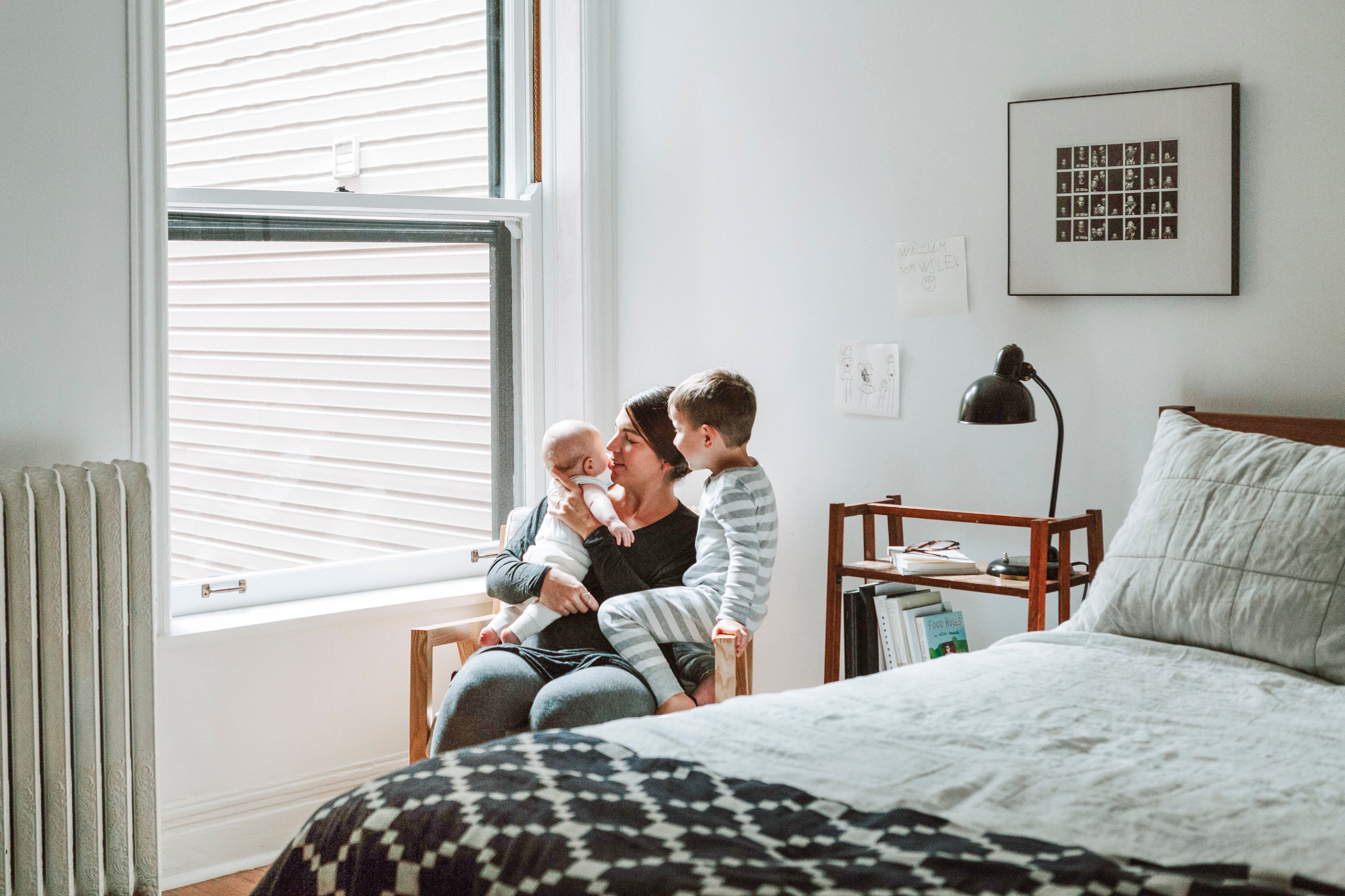 person holding a baby and a child sit on a chair next to a bed