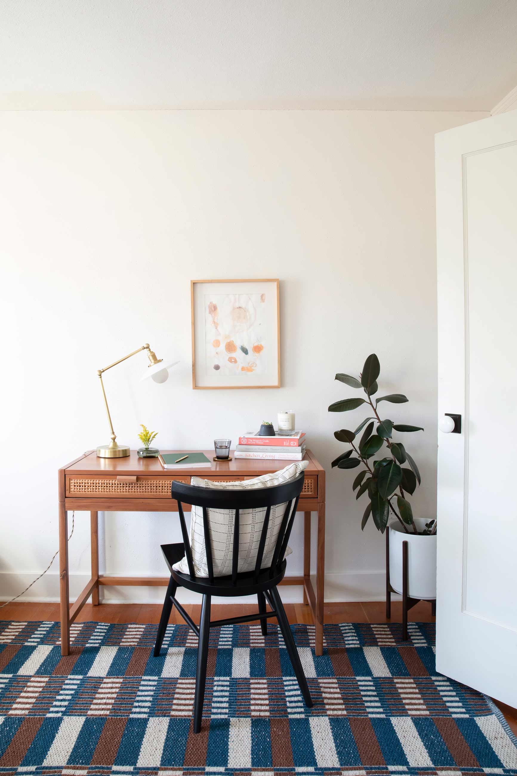 Office desk with table lamp and books atop it, with a rubber tree plant next to it and a blue and brown wool rug beneath it. 