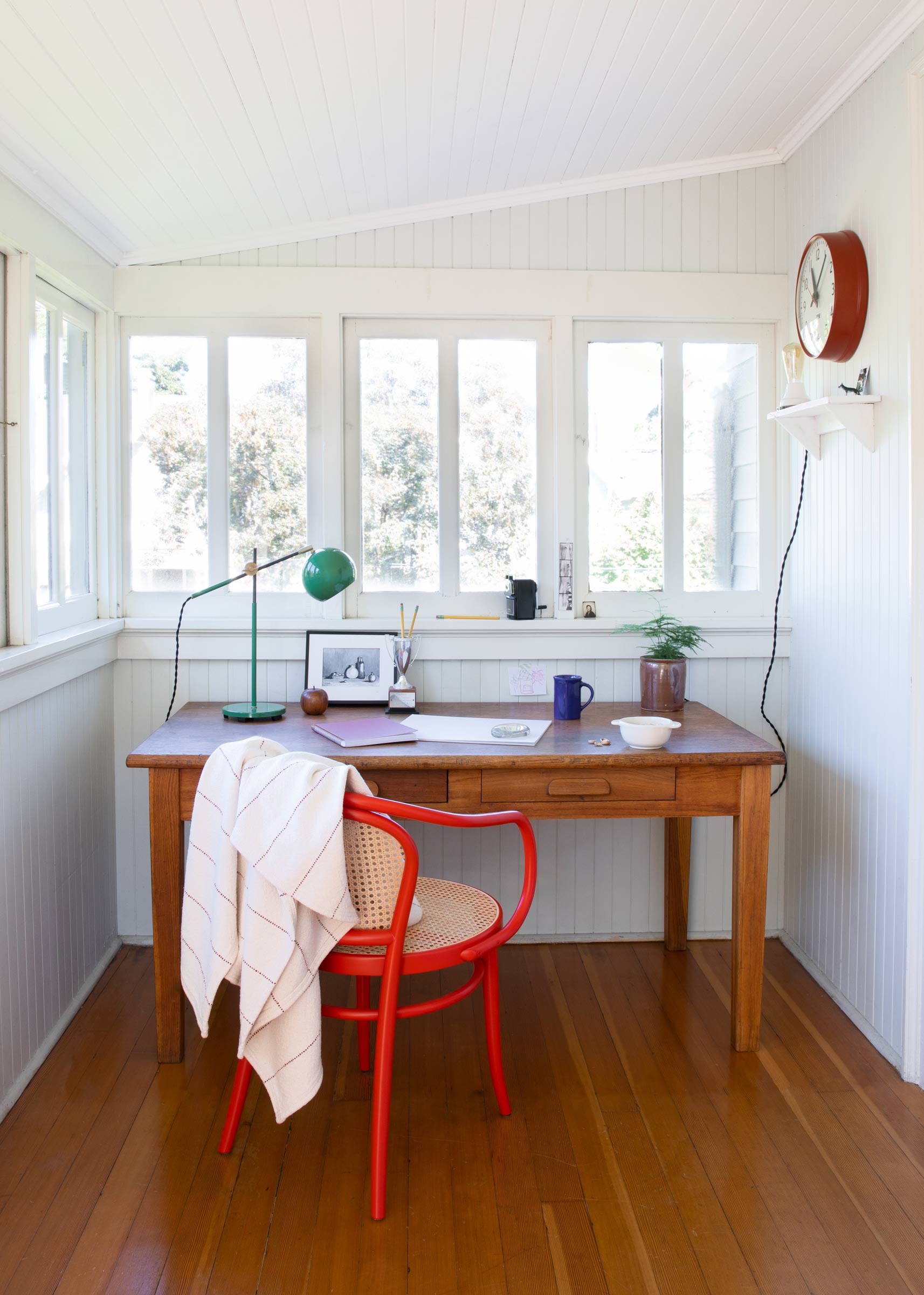 Wooden desk in a bright office with a red wall clock, green desk lamp, and red cane chair. 