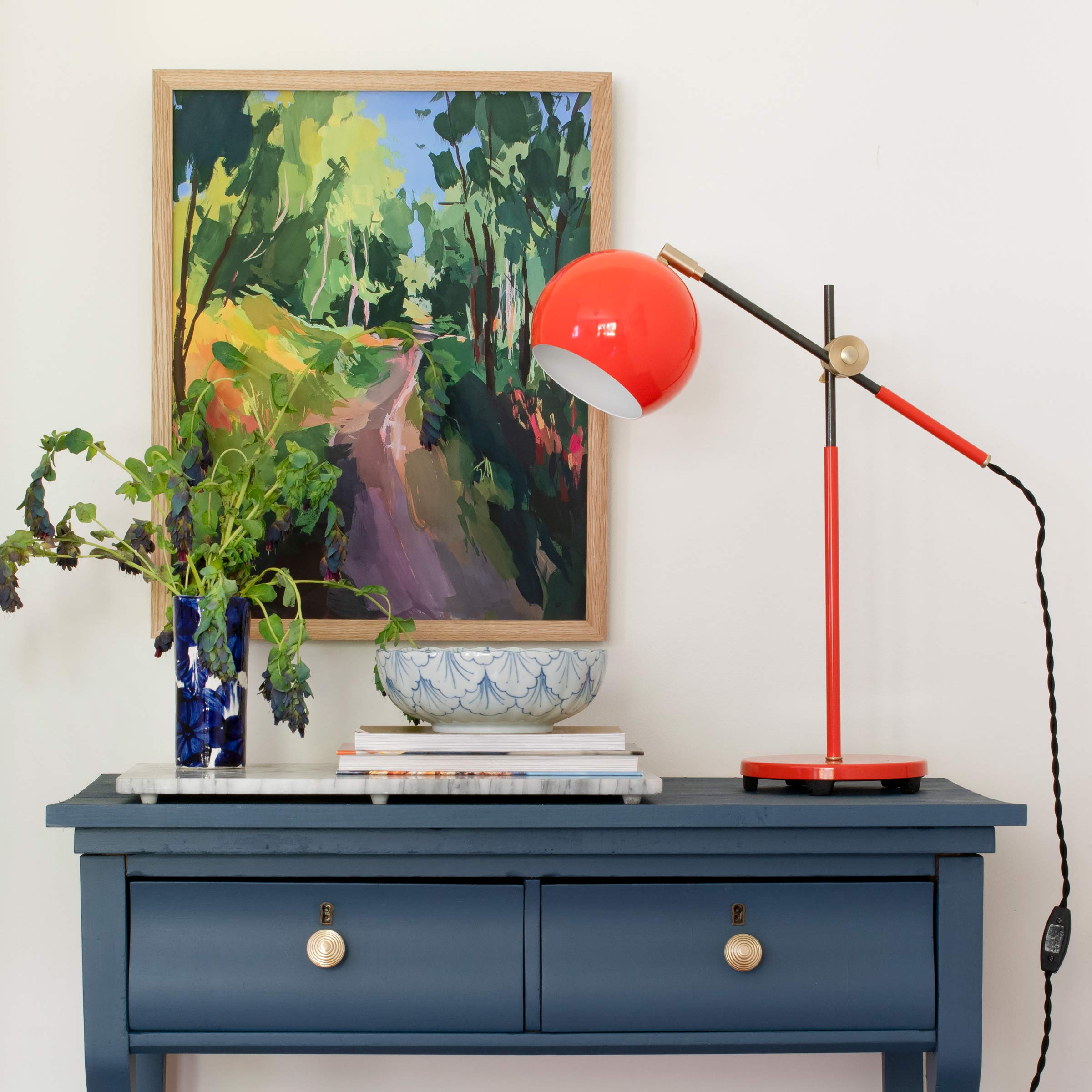 Original framed art above a blue side table with a bowl, vase, and red lamp on top.