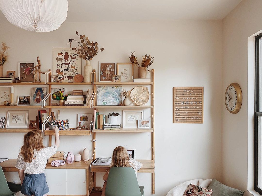 couple of girls sitting in a room with a clock on the wall and shelves