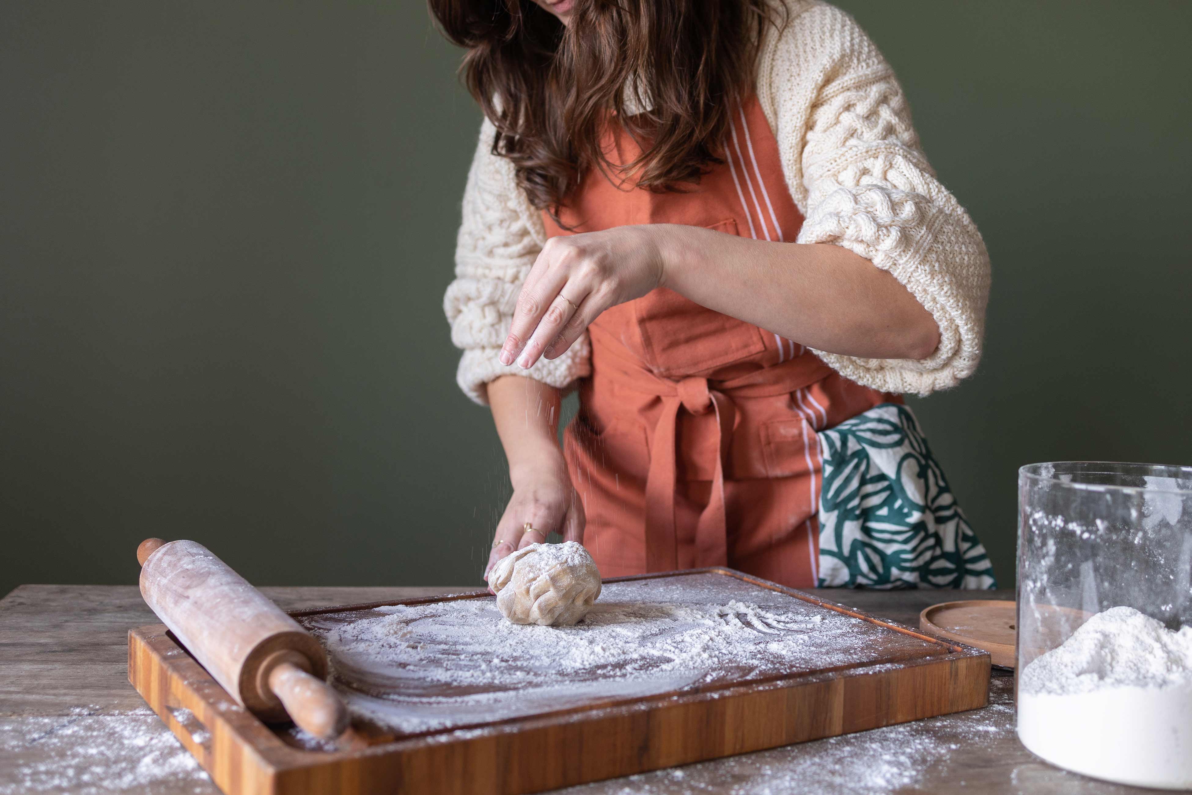 Person sprinkling flour over ball of gingerbread dough on top of butcher block cutting board. 