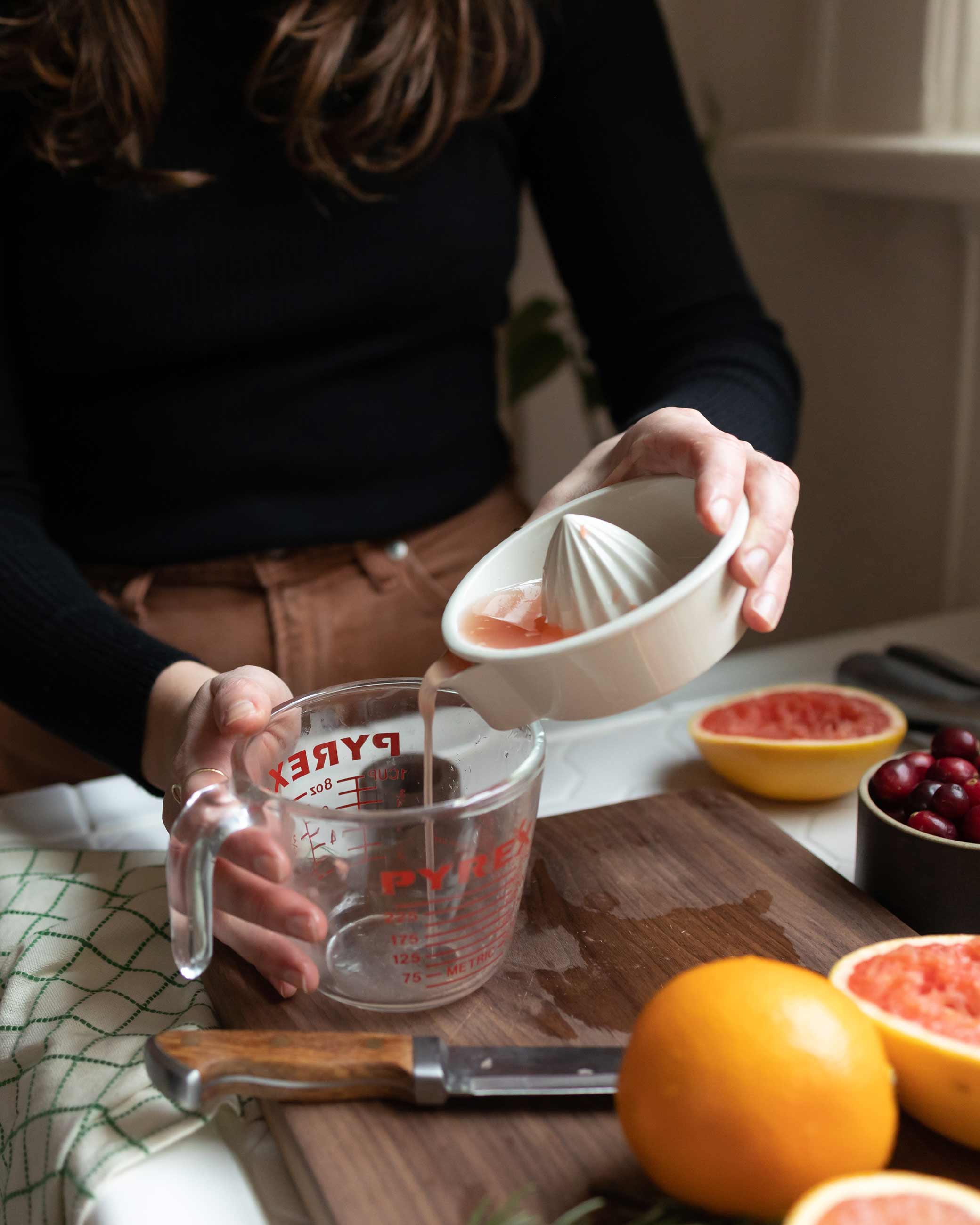 Person straining grapefruit juice into measuring cup on wood cutting board.