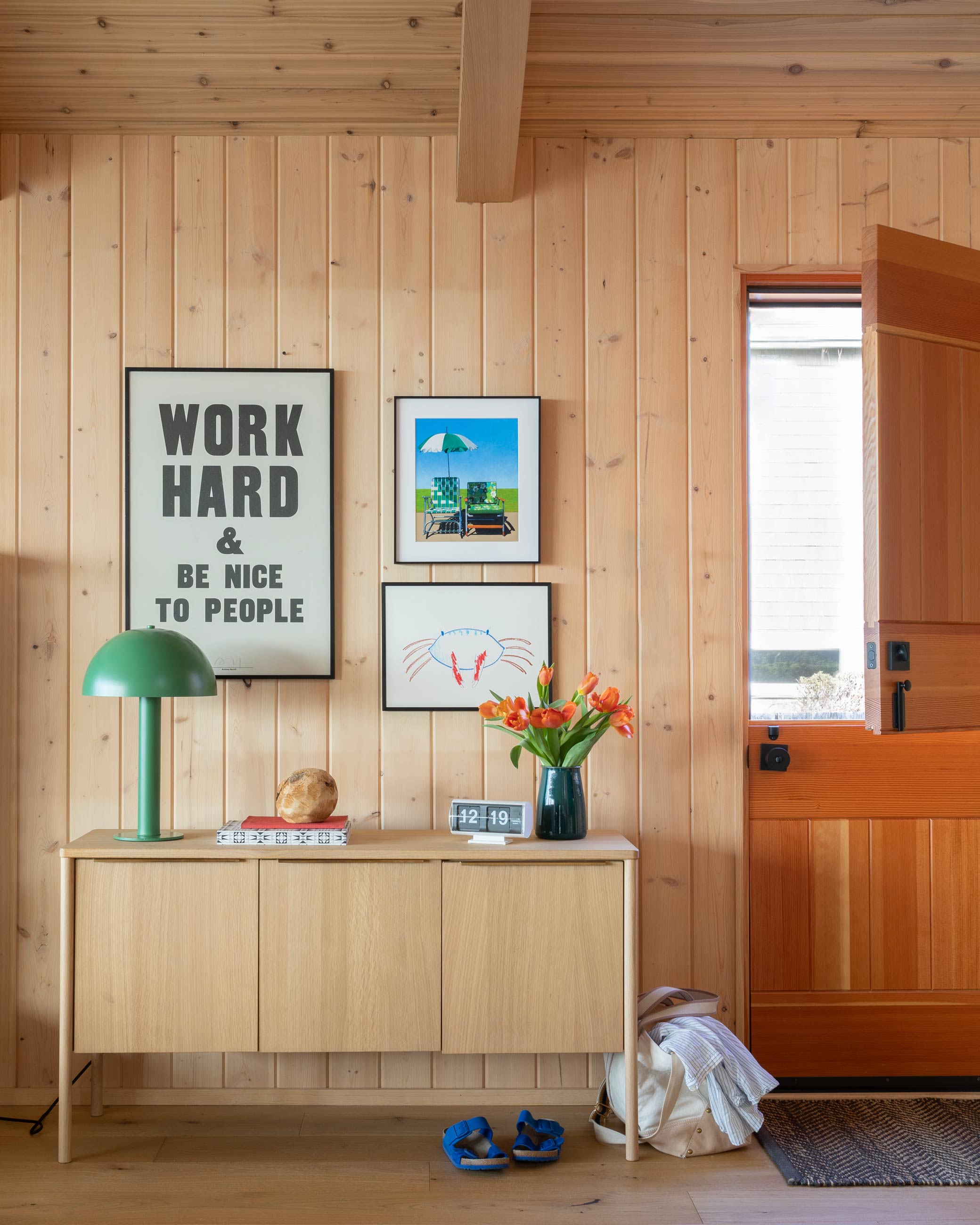 Green lamp sitting atop oak sideboard next to door with various art prints above. 