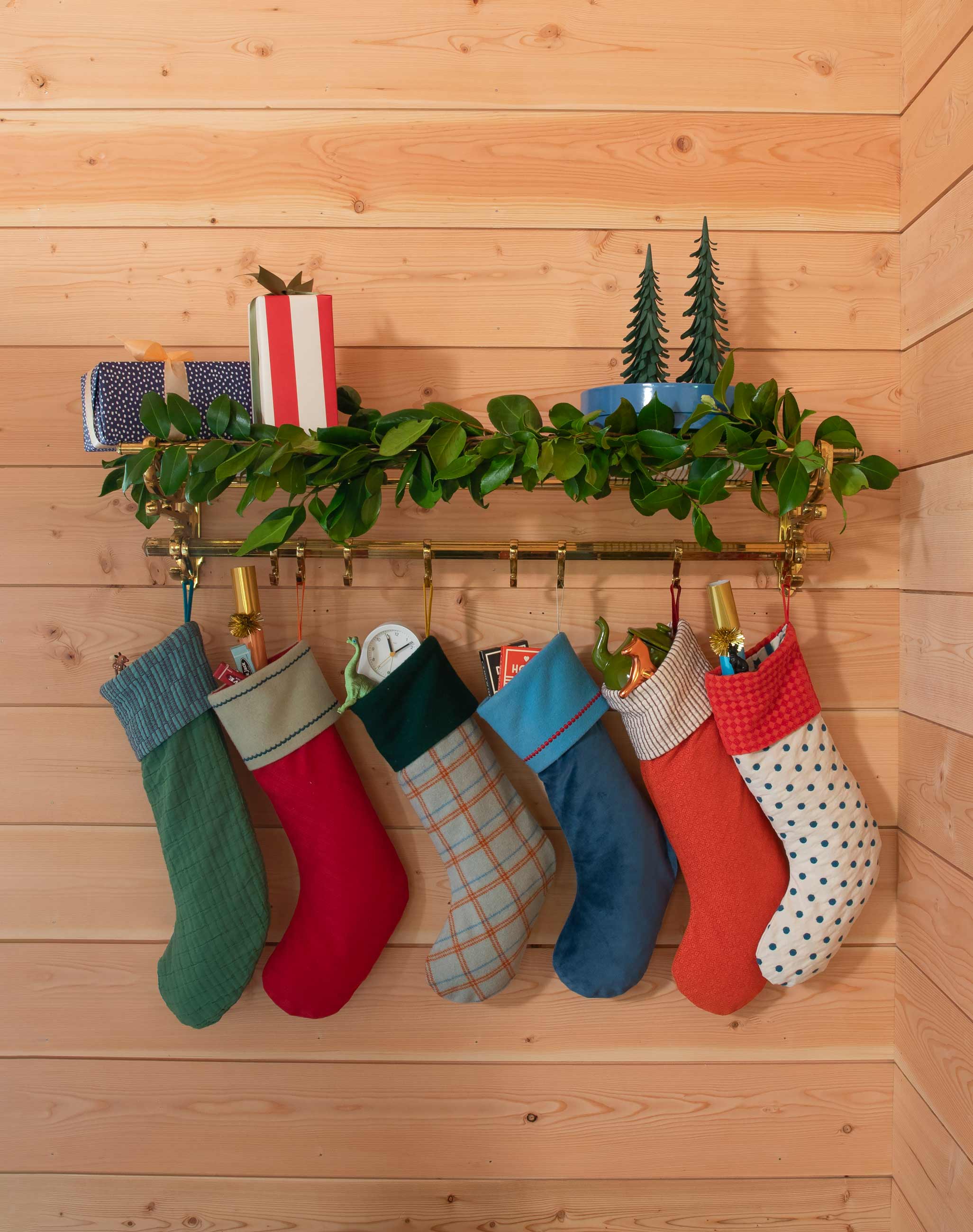 Colorful stockings hanging on gold peg rail with shelf above holding greenery and holiday decor.