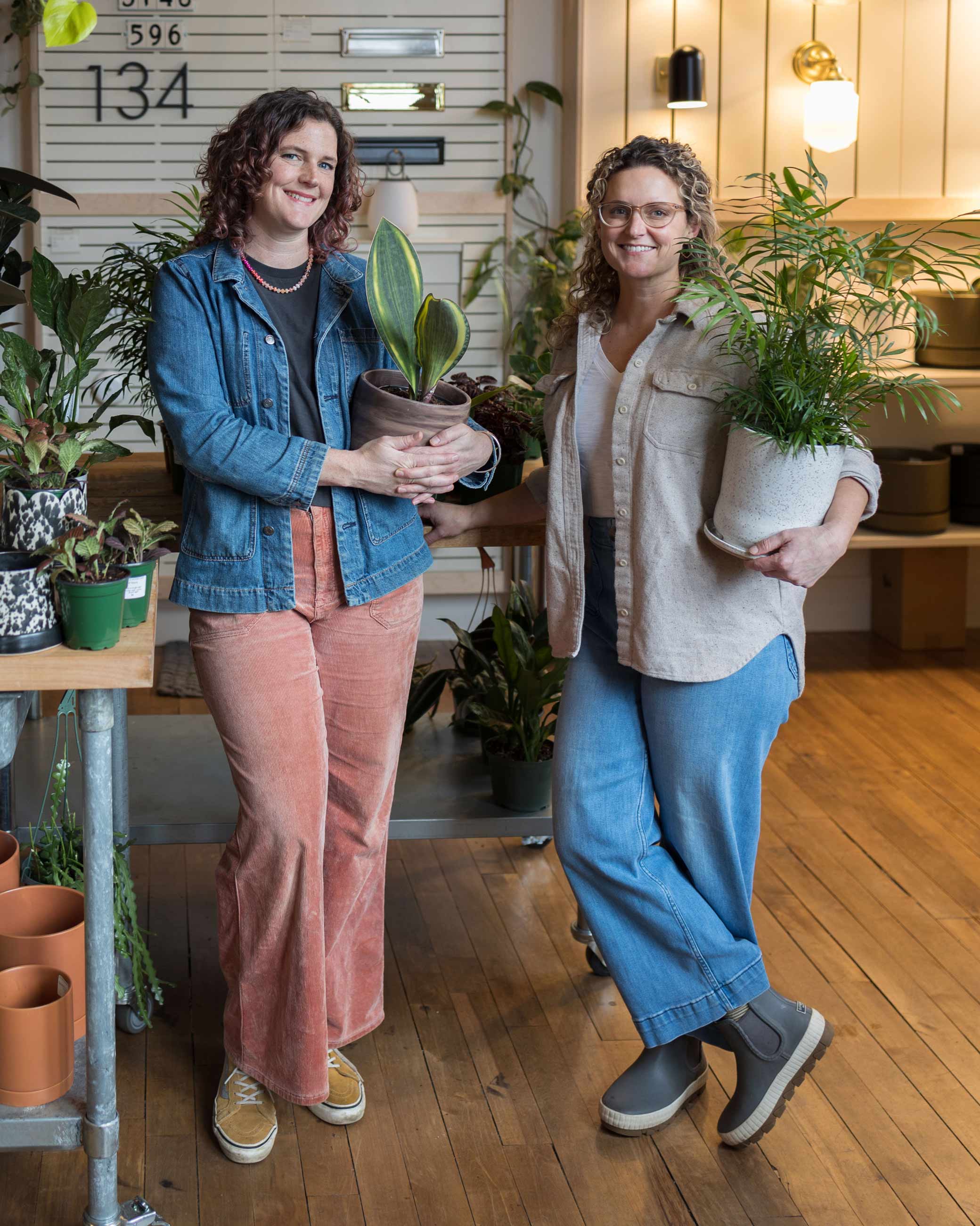 Two sisters standing in plant shop holding potted plants. 