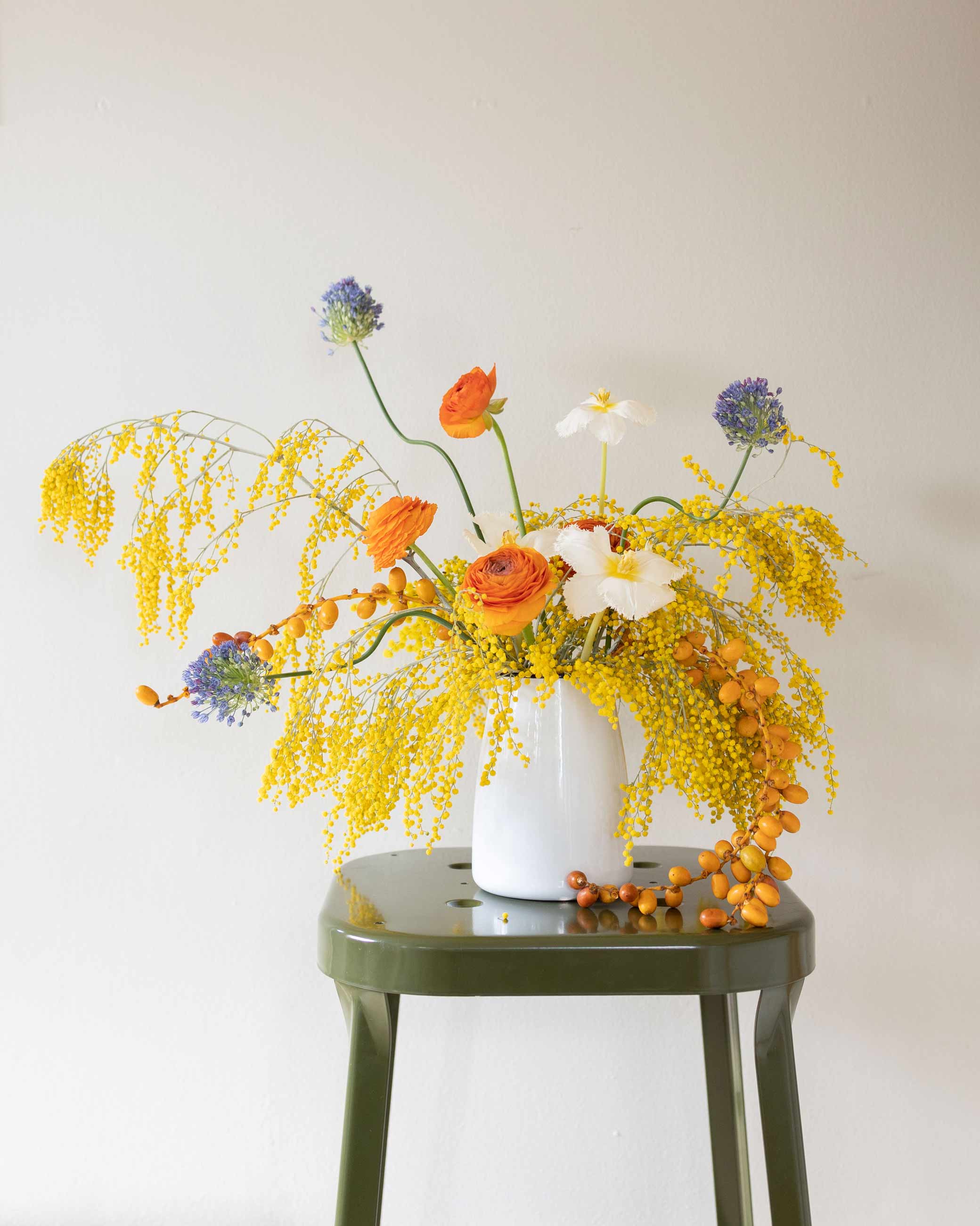 Vibrant floral arrangement in enamel crock sitting on green stool. 