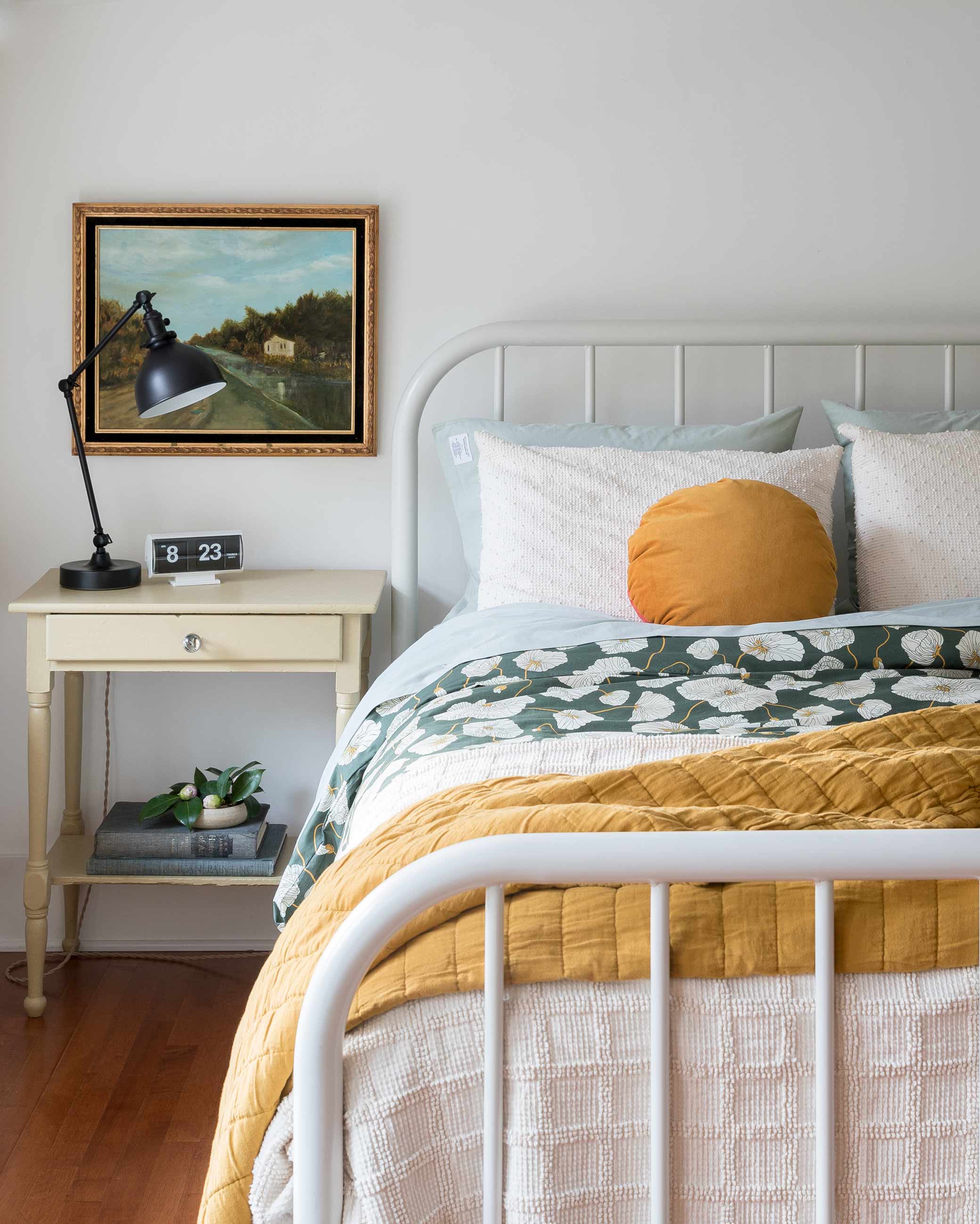 Floral duvet and yellow quilt on white bed frame with a vintage side table and lamp