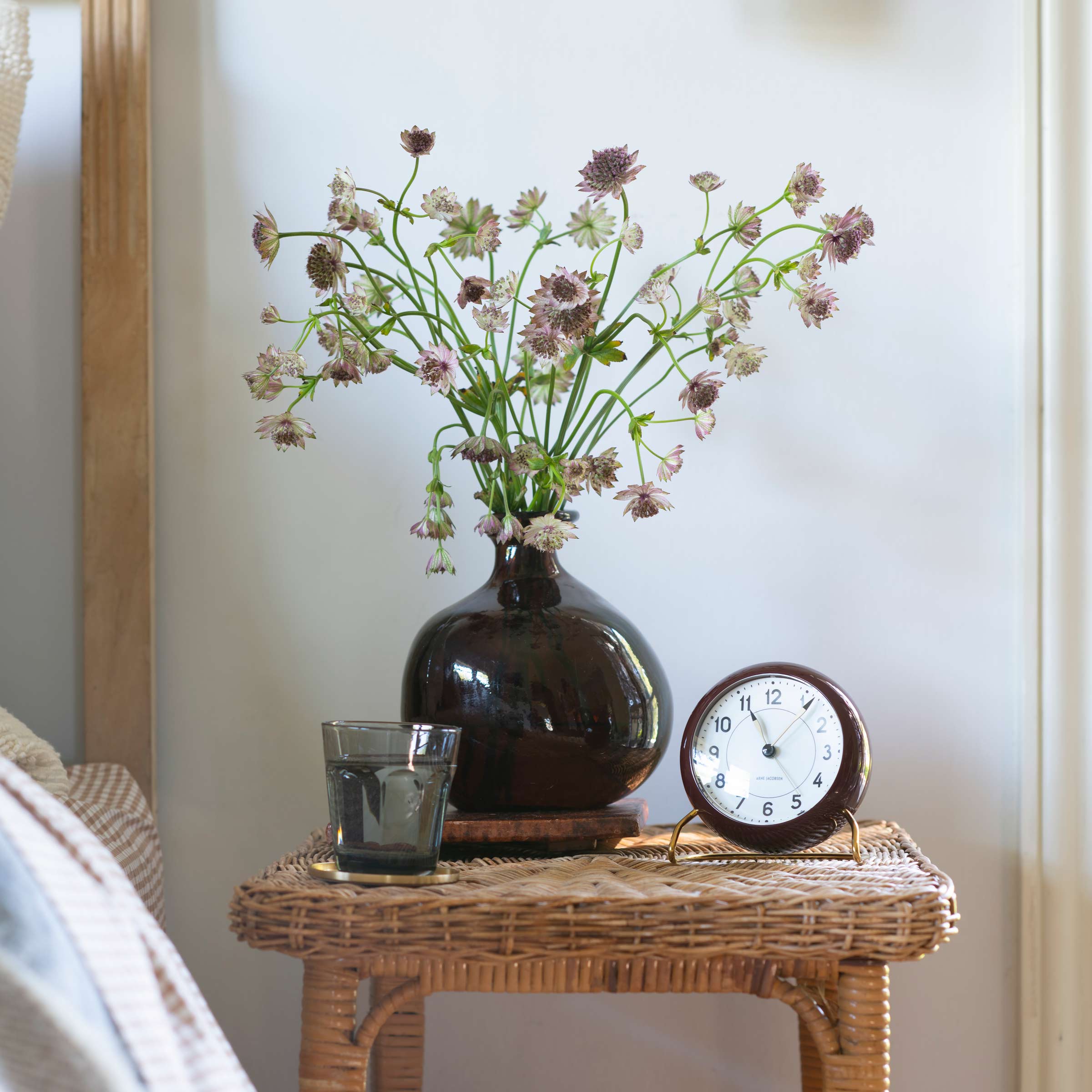Wicker side table with flowers in a vase, glass of water, and a burgundy alarm clock.