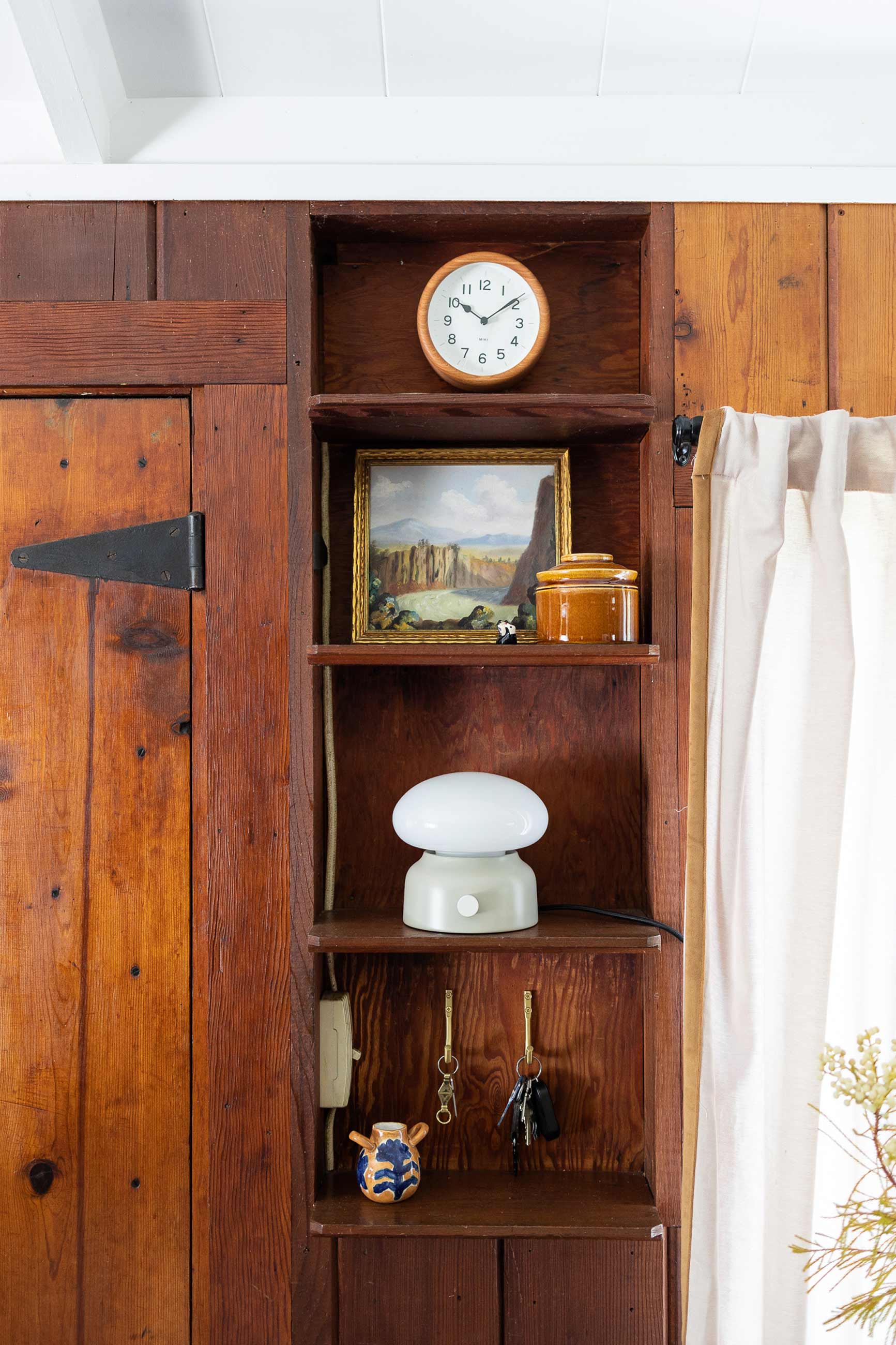 Wooden alarm clock sitting in built in wooden shelf above lamp and key hooks.