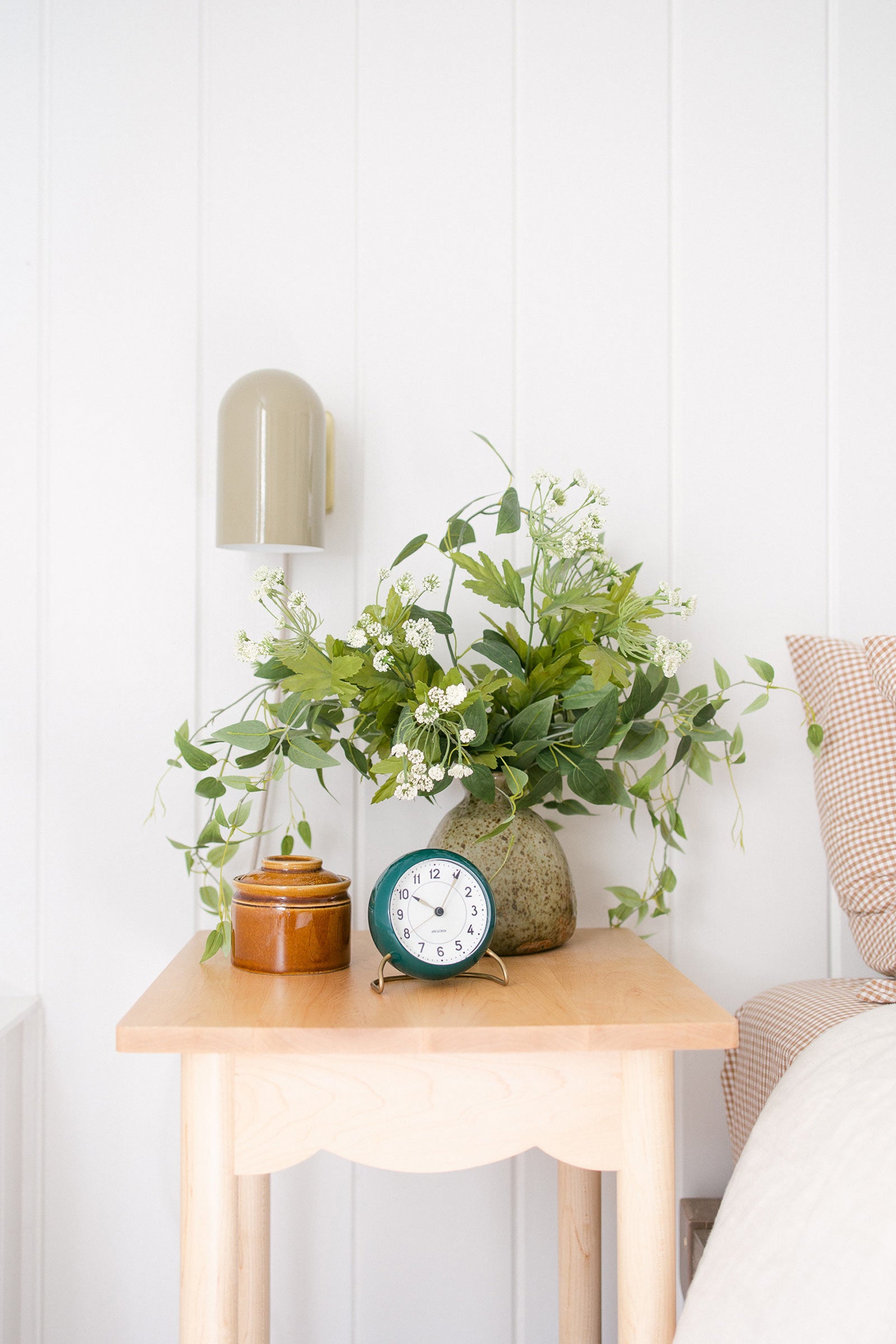 a wooden scalloped side table with lush greenery and small blue clock and auburn ceramic jar.
