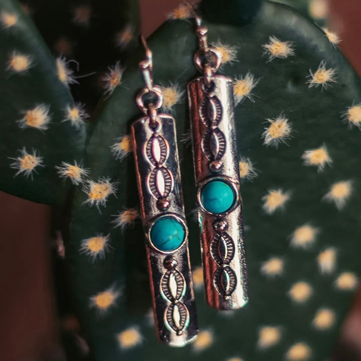 The Outlaw Stick Earrings are a 2" silver stick dangle with a small turquoise stone and stamped oval design. The earrings are on a fish hook back. The earrings are super lighweight. 