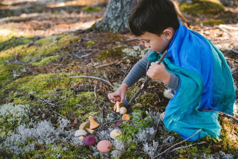 Boy playing with wooden mushrooms in mossy forest wearing playsilk