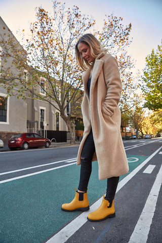 Woman crossing the road in a pair of Mustard colour Bobbi Gumboots by Merry People