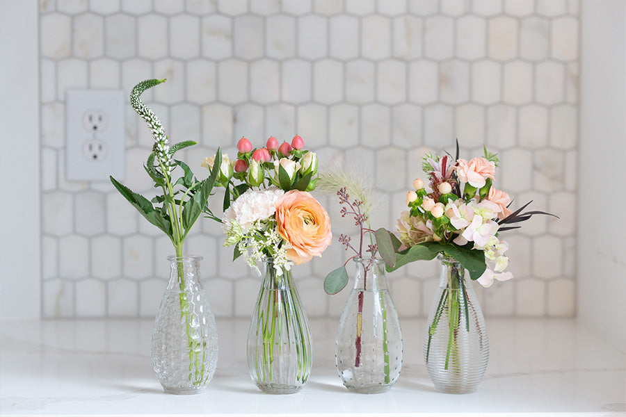 Four glass bud vases wit peach and white flowers in them.