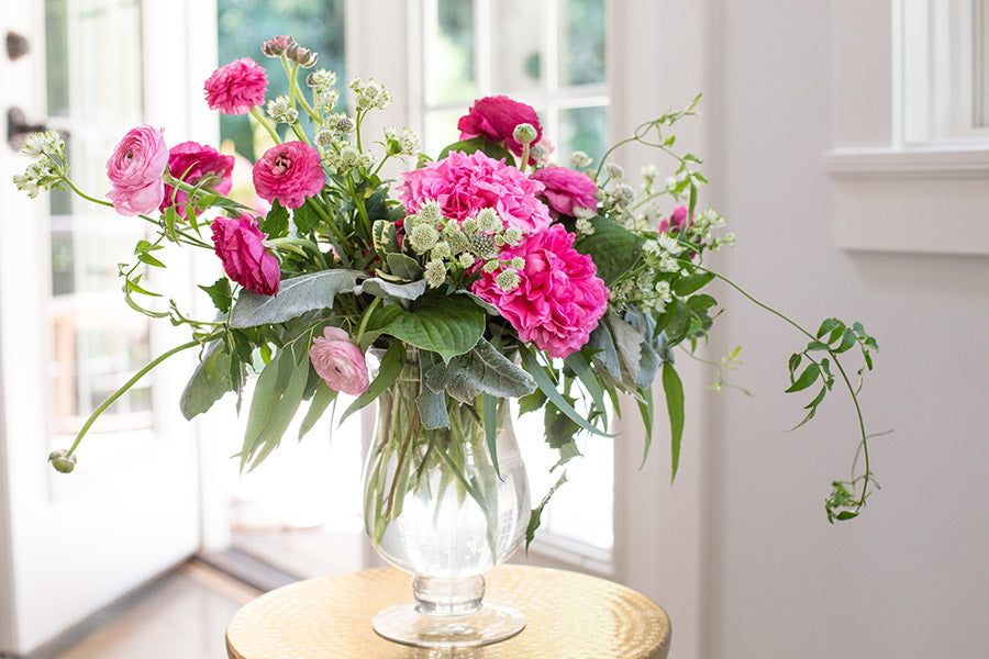 A clear vase with bright pink flowers and a variety of wide leaf and small leaf greenery