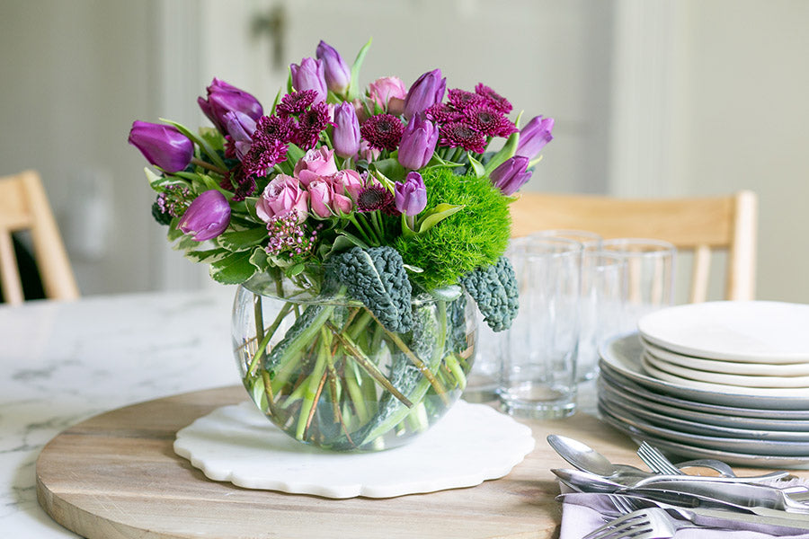 A purple and green flower arrangement in a round glass vase 