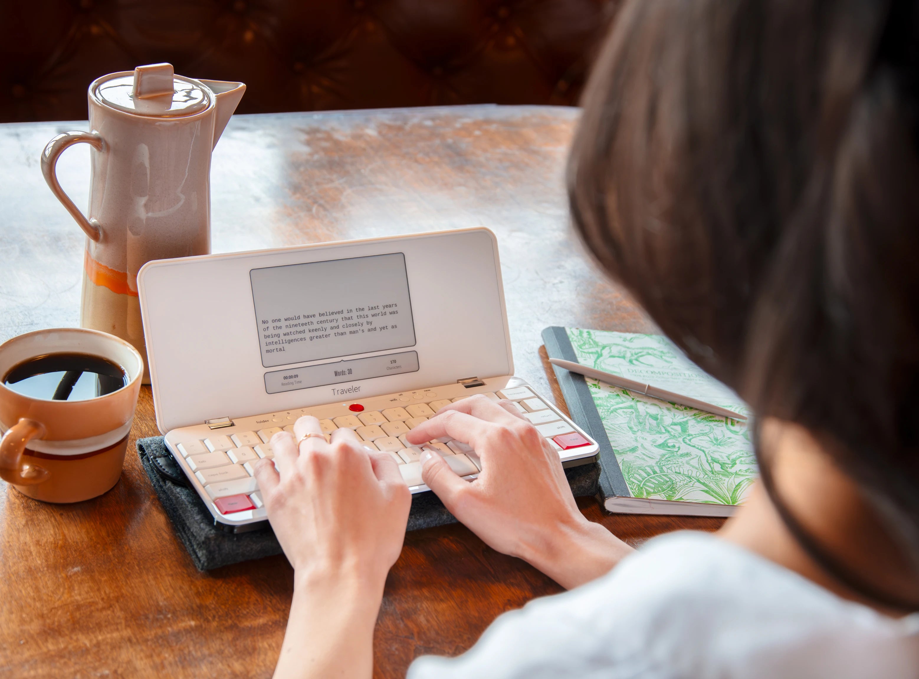 Person typing on a small portable typewriter with coffee and a notebook nearby.