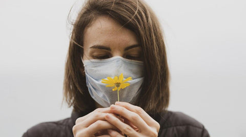 mujer castaña con mascarilla celeste pálido oliendo flor amarilla