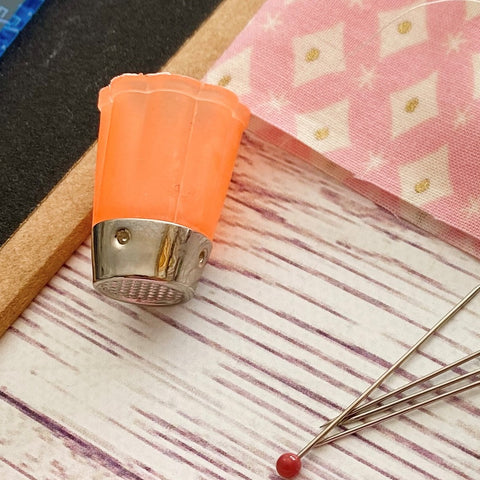 orange silicone thimble with a metal tip sits on a painted timber board. In the background a snipped of pink fabric is showing and pins are in the forground