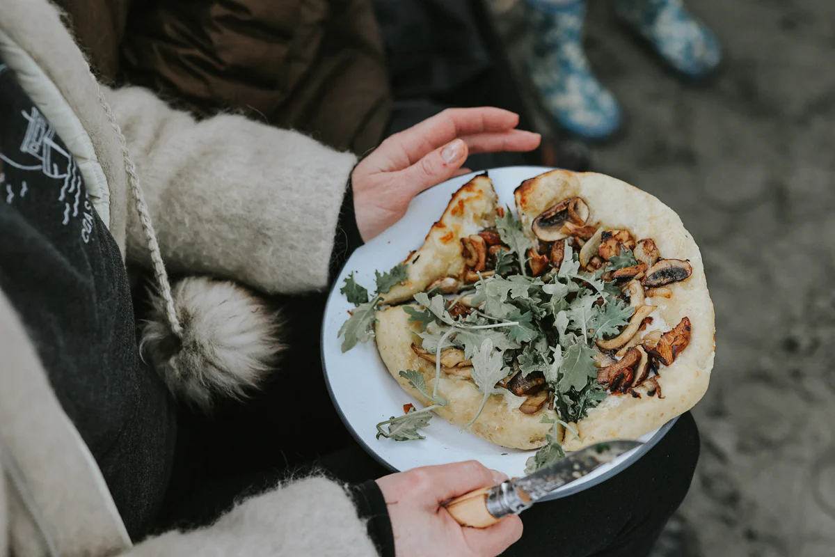 Woman holding pizza on beach with falcon enamelware plate and opinel knife