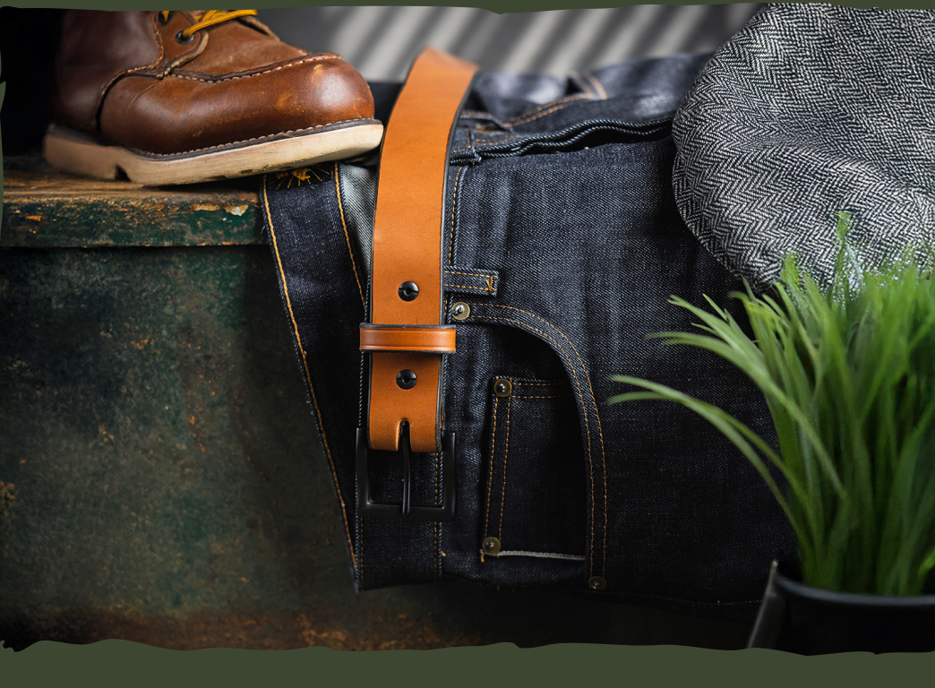 Brown shoe, leather belt, and denim jeans with a hat on a rustic surface.