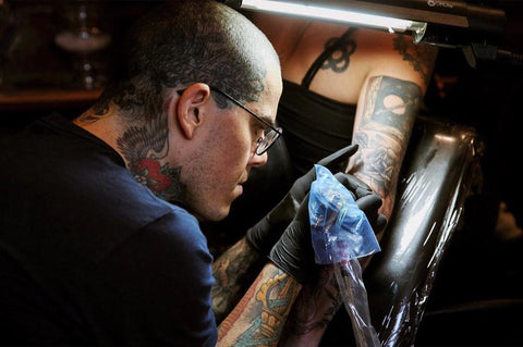 Ryan Roi - Tattoo Artist and Financial Coach for Creative Professionals - The Artful Dollar Business Coaching - Pictured Ryan Roi Focusing on Tattooing a Client's Elbow Tattoo