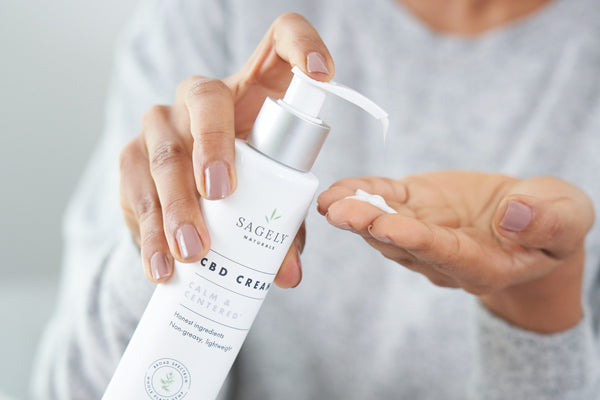 A close up image of a woman holding a bottle of Sagely Naturals Calm & Centered CBD Cream as she presses some out into her hand.