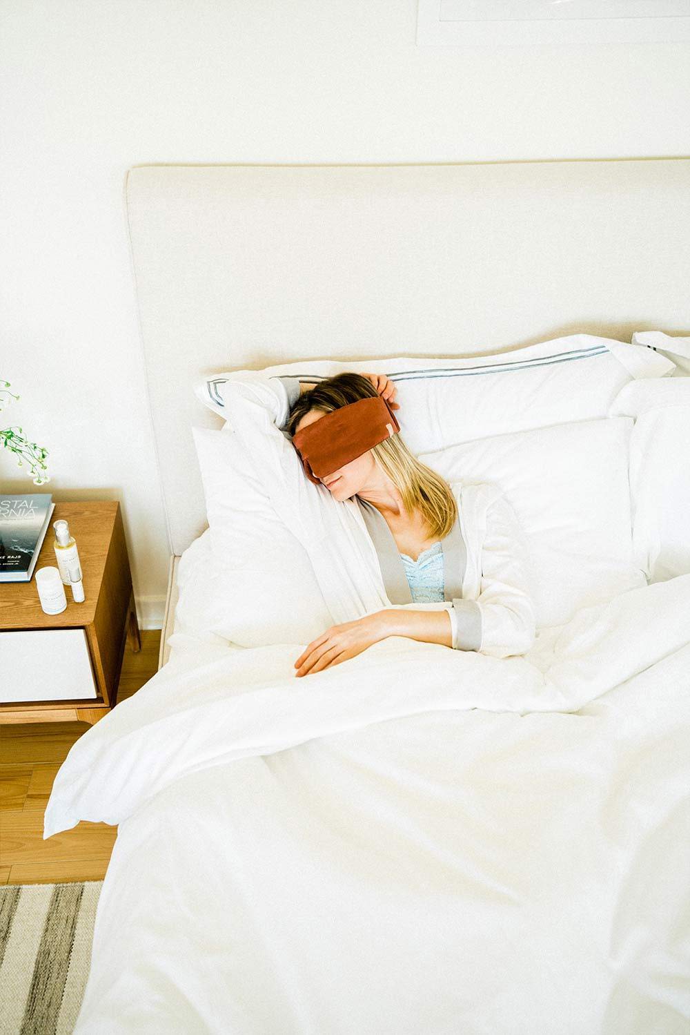 Woman lying in bed with white sheets and eye mask on and a bedside table with Sagely Naturals products.