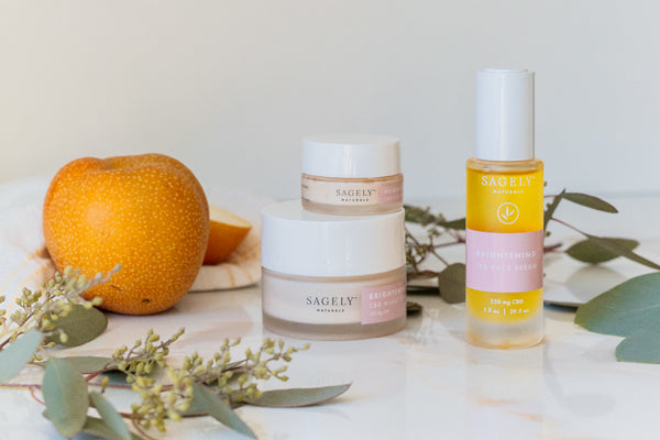 A picture of Sagely Naturals Brightening CBD Skincare collection and a pear on top of a marbled tabletop with decorative greenery throughout.