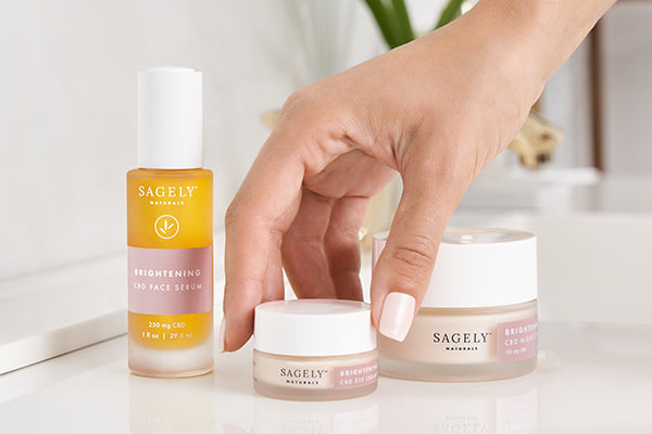 A hand picking up Sagely Naturals Brightening CBD Eye Cream which is sitting on a sink counter next to CBD Serum and CBD Night Cream