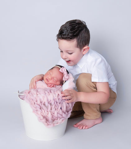 A young boy in a white shirt kneels beside a white bucket prop, gently cradling a sleeping newborn girl wrapped in a pink blanket with a pink bow on her head, expressing a tender moment of sibling connection.