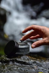 womans hand reaching into touch charcoal facial mask in front of a waterfal