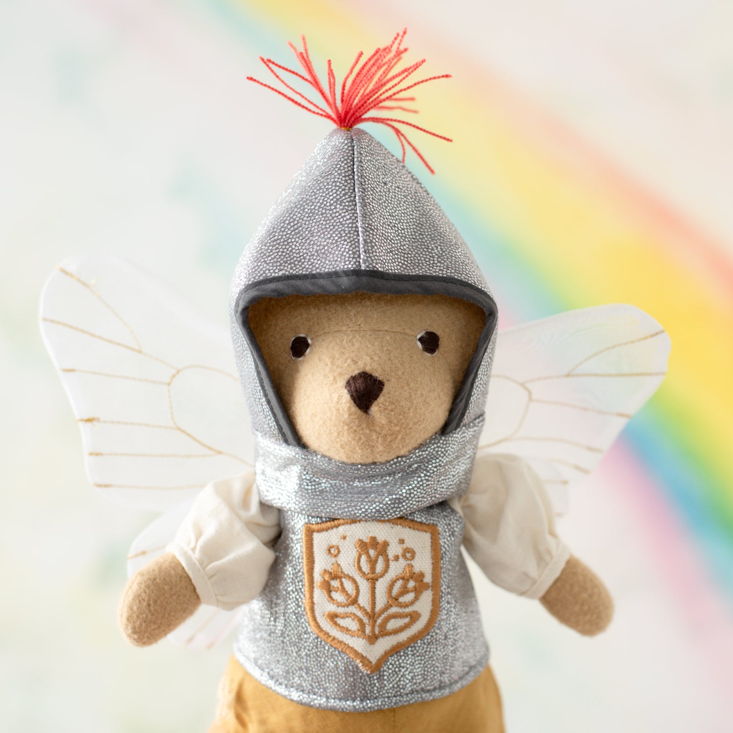 Portrait of Nicholas Bear Cub wearing shiny gray doll helmet with red plume, shiny gray vest with gold shield on the front, a poofy sleeved white shirt, and fairy wings. He is in front of a rainbow backdrop.