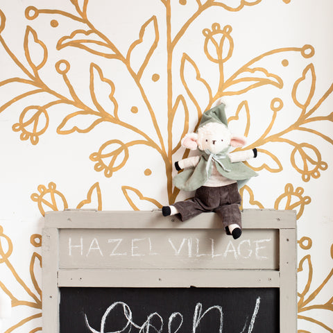 Arthur Lamb sits in his hooded cloak/capelet in front of a golden floral backdrop. He sits on top of a sign that says "HAZEL VILLAGE Open!"