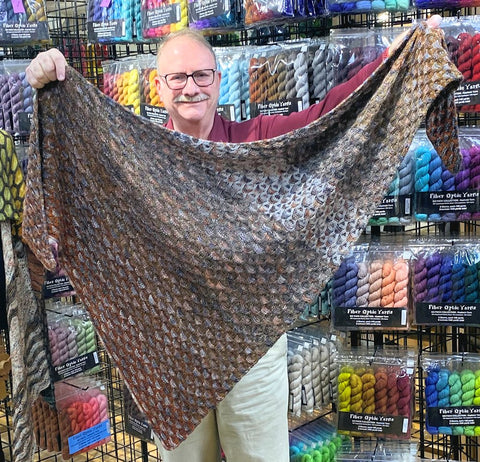 A man holding a triangular shawl showing the bands of a Fiber Optic Yarns six pack fade set progressing in towards the middle and then back out towards the other edge.